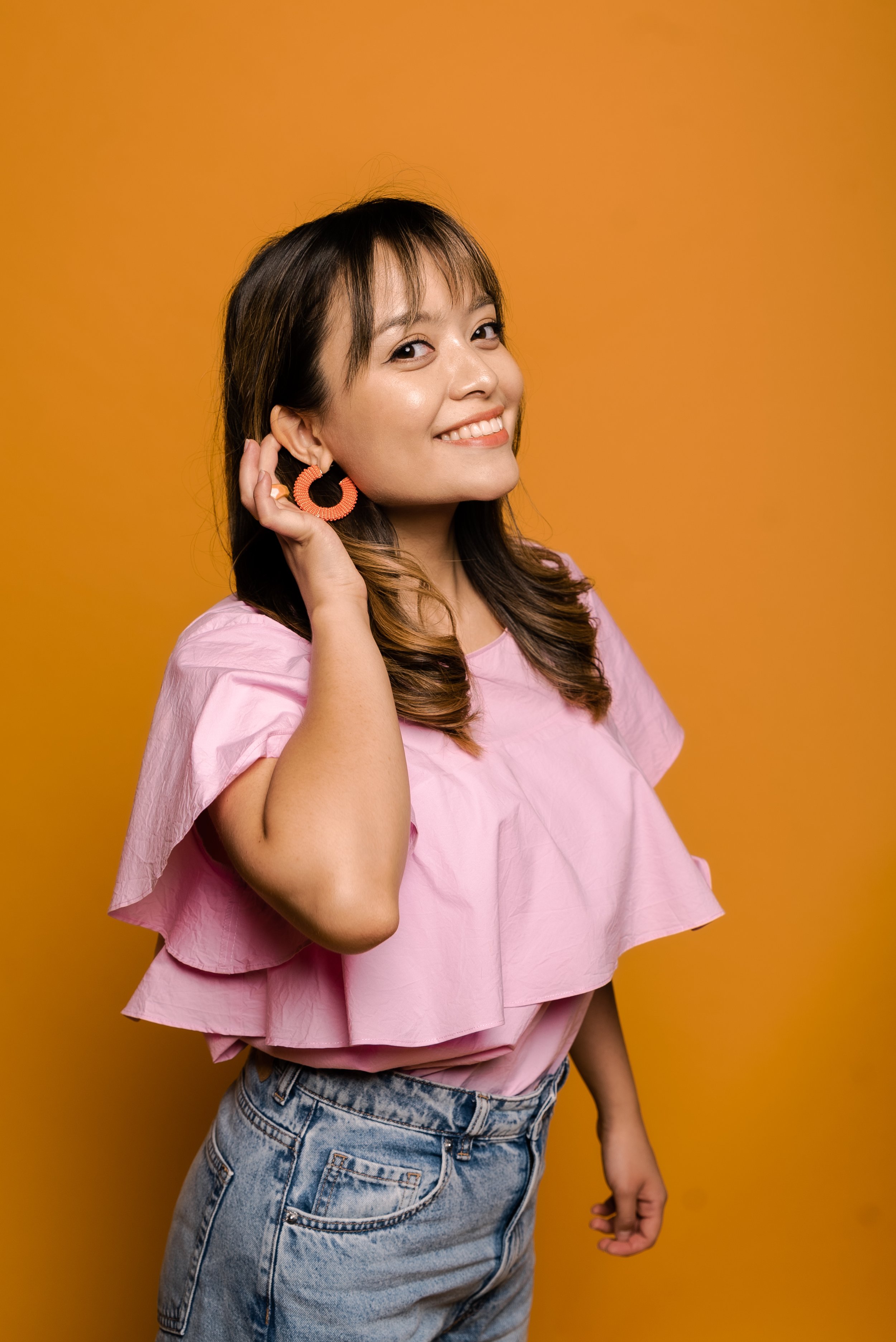 Woman smiling in a pink top and blue jeans with orange circular earrings against an orange background.