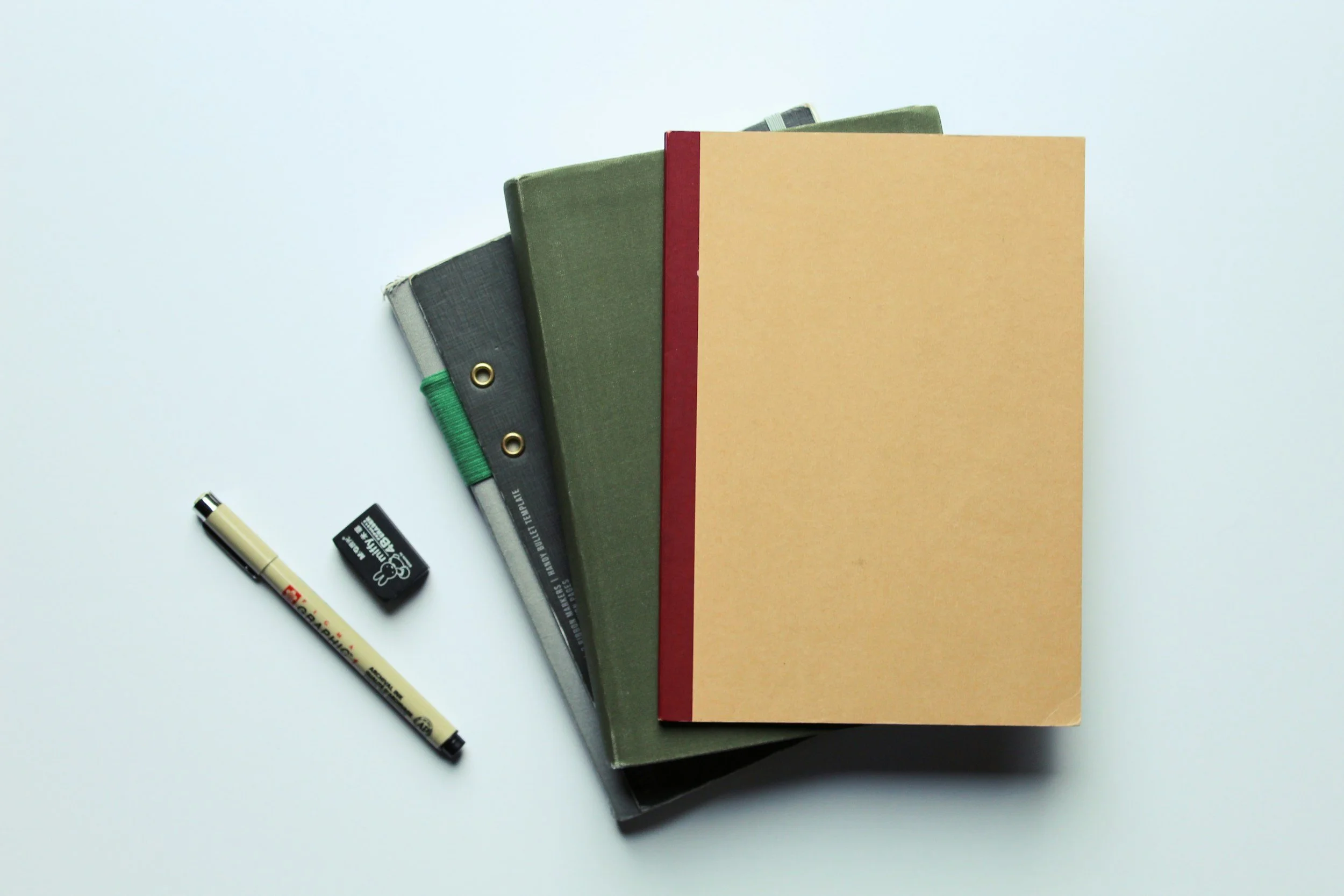 A stack of three notebooks in different colors - black, green, and beige - with a red spine, on a white surface, with a black pen and black eraser nearby.