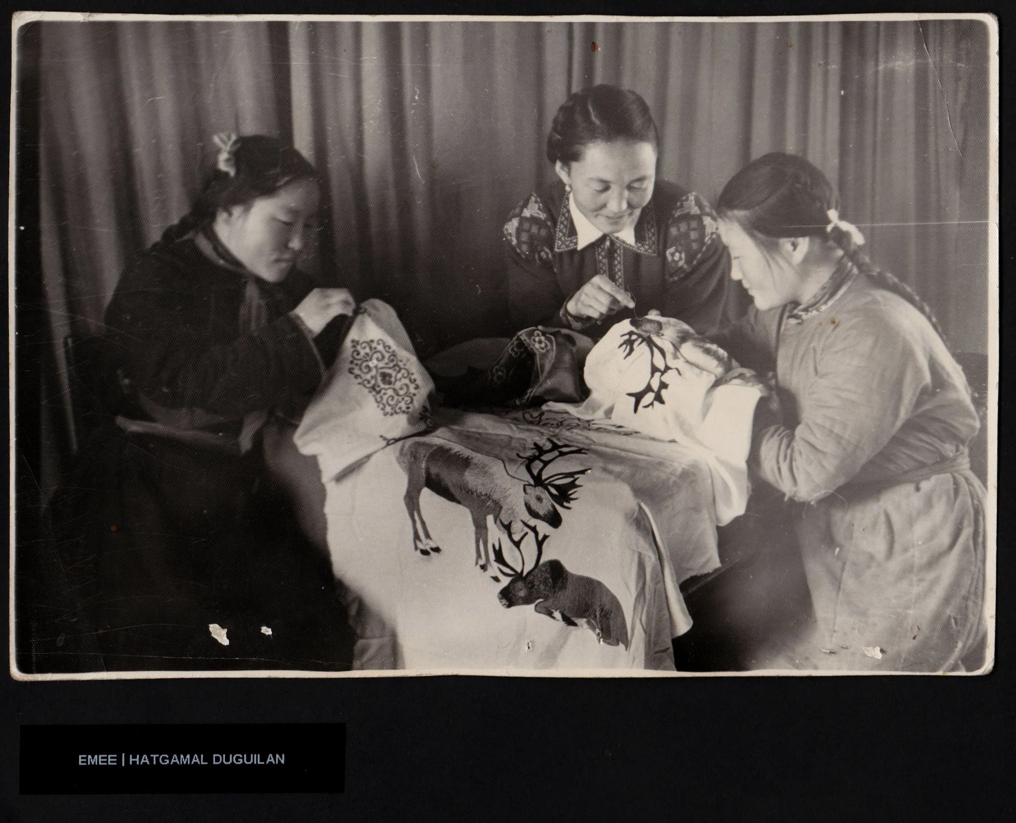Black & white photo of three Mongolian women with traditional embroidered clothing on a table covered with a cloth featuring reindeer & animal embroider. The women are focused on teacher, possibly performing a traditional craft or care activity.