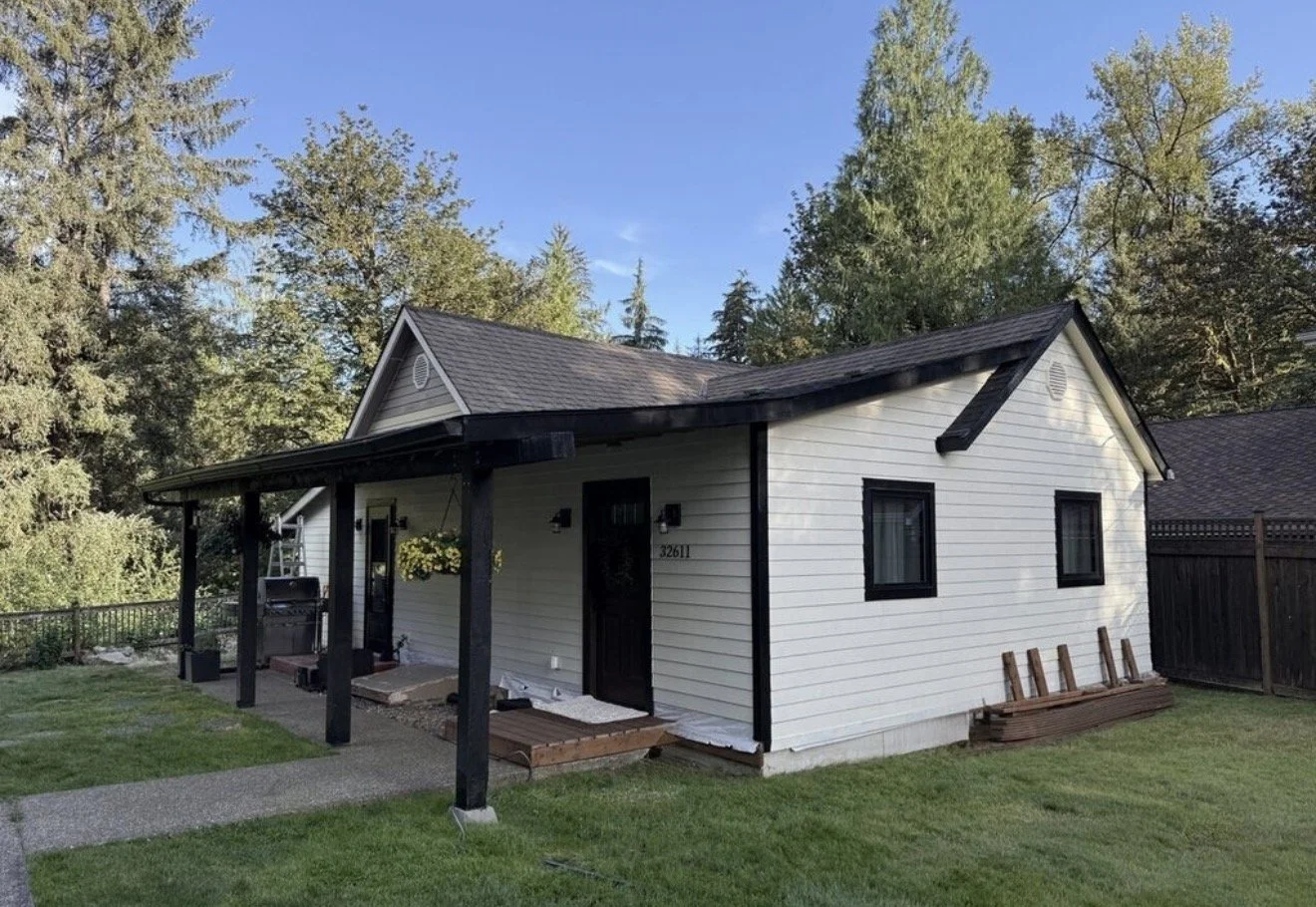 A small white house with black trim and a gabled roof, featuring a covered porch with black support beams, situated in a backyard with green grass and surrounded by trees.