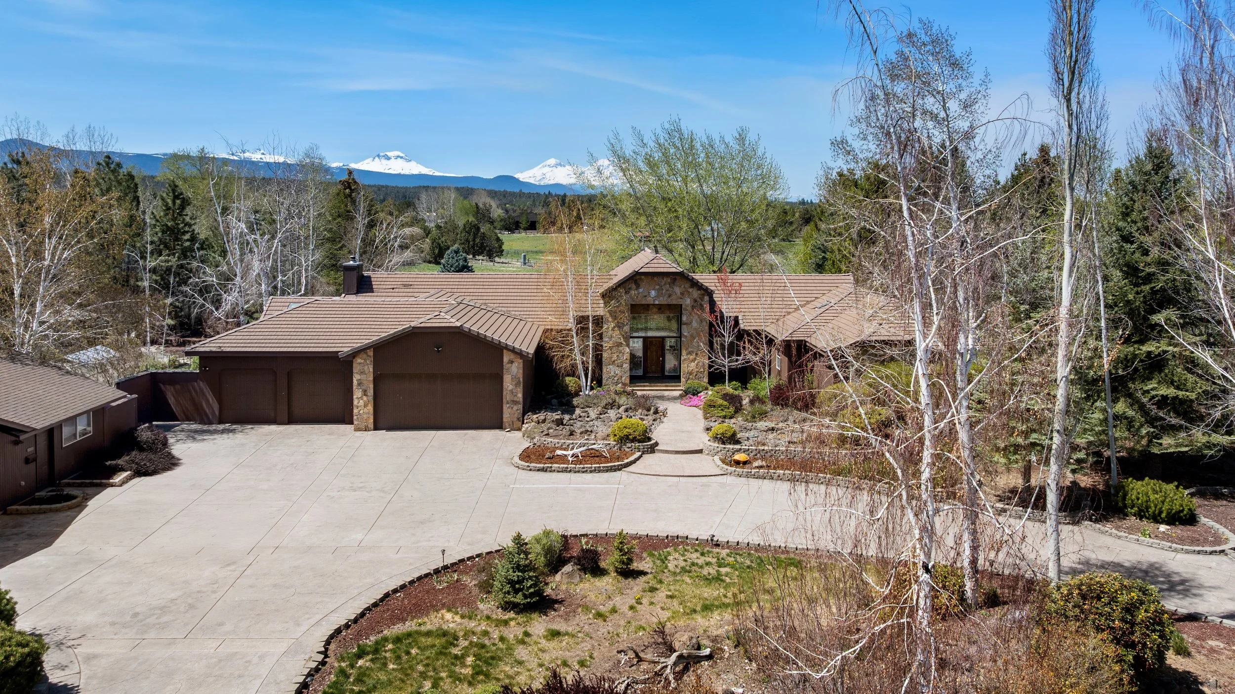 A modern house with a stone facade and brown roof surrounded by trees, with snow-capped mountains in the distance under a clear blue sky.