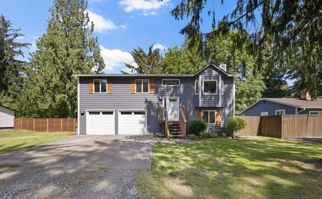 A two-story gray house with white garage doors, wooden shutters, and a small front porch with stairs and a white door, surrounded by green trees and lawn.