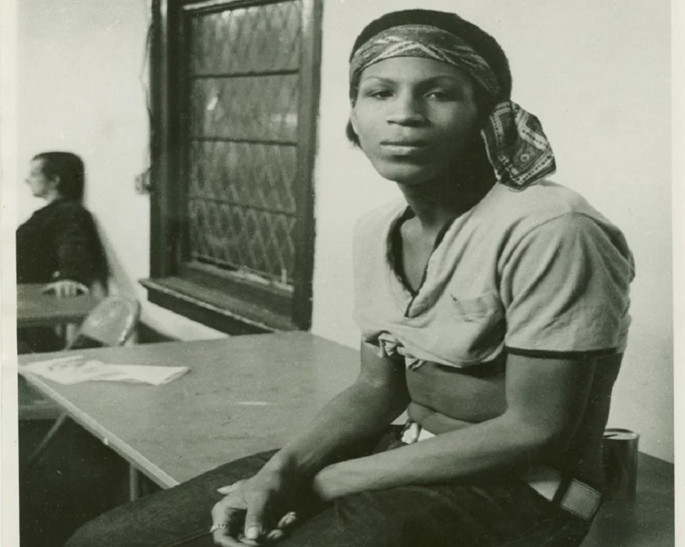 A young black woman sits on a folding table. She wears a bandana tied around her forehead with a crop top and jeans.