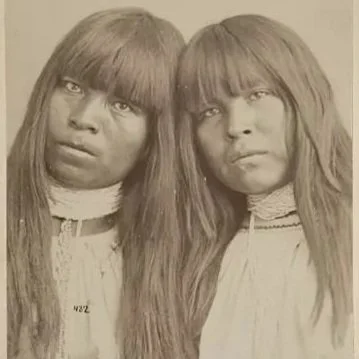 Two people with long wavy hair and bangs lean into each other. They are both wearing a white garment and a lacy white neck adornment
