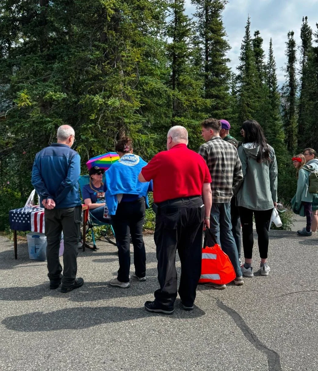 seated ranger under rainbow umbrella speaks to individuals gathered around her.