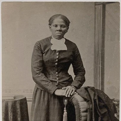Black and white photo of a black woman in a dark 19th century dress with a white collar. Her hands are folded and placed on a chair back.