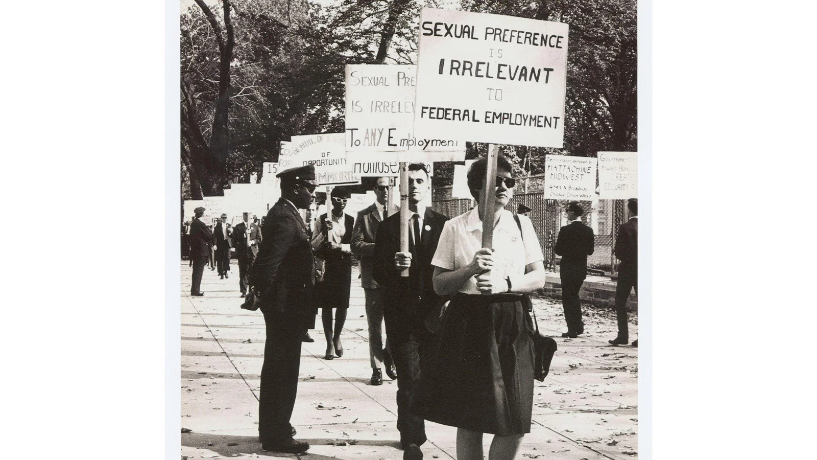 A line of protesters hold signs on picket. The woman at the front of the line holds one that reads "Sexual Preference is Irrelevant to Federal Employment"
