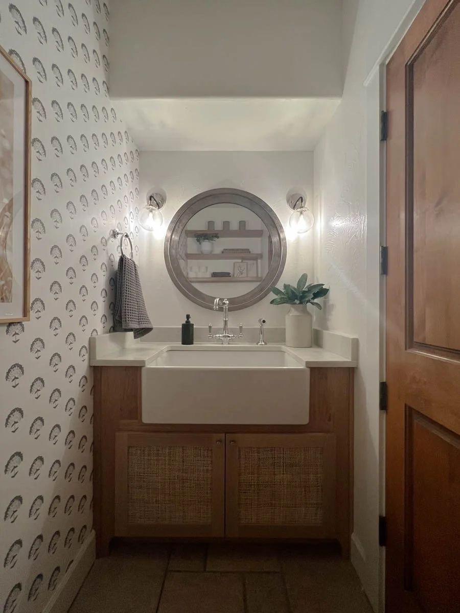 Bathroom with patterned wallpaper, round mirror, and custom vanity by Verity Construction.