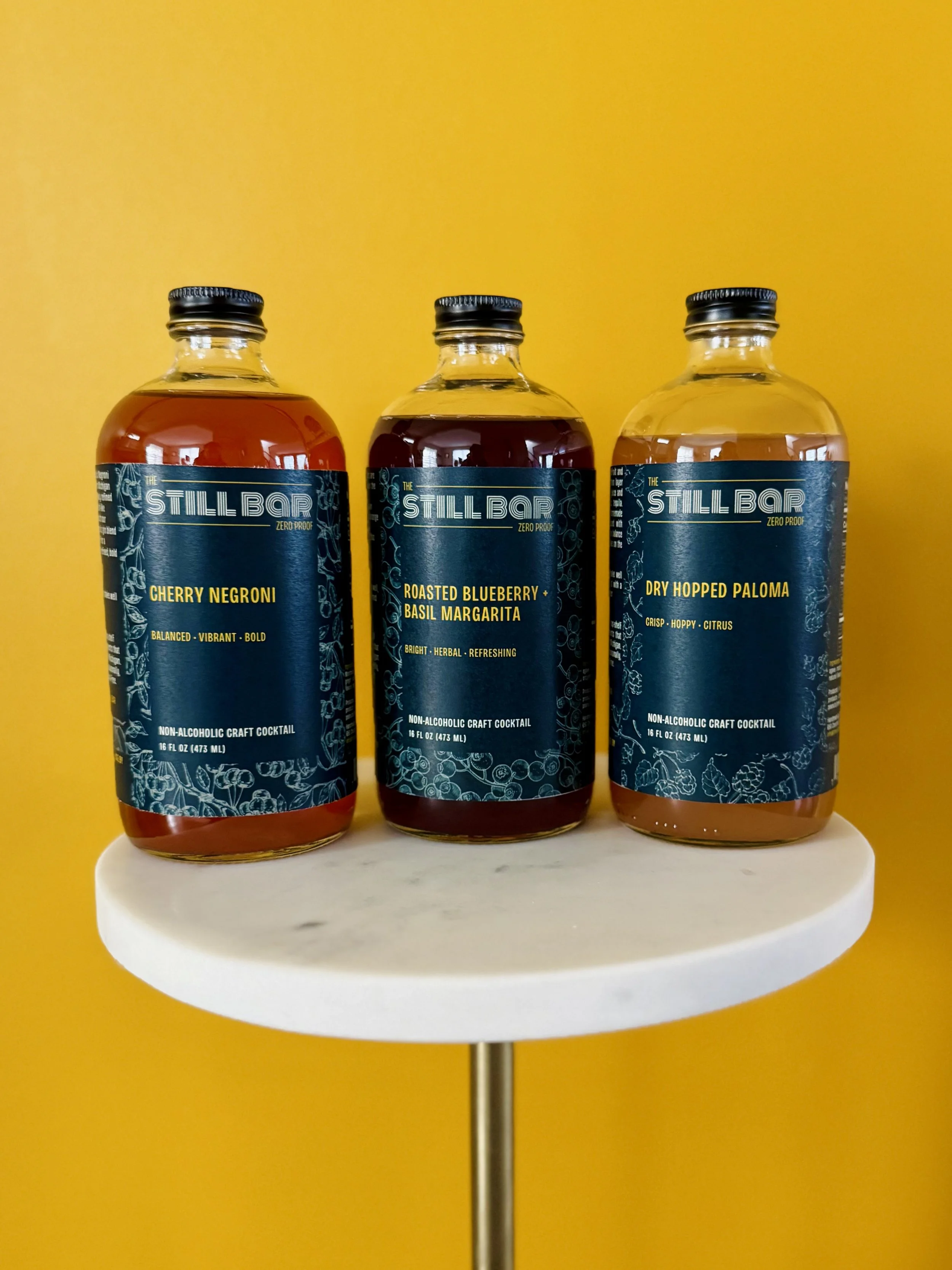 Three bottles of Still Bar non-alcoholic craft cocktails on a white marble-topped table against a yellow background. Flavors from left to right: Cherry Negroni, Roasted Blueberry + Basil Margarita, Dry Hopped Paloma.