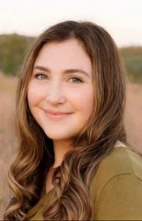 A young woman with long wavy brown hair and a light smile, wearing an olive green top, outdoors with blurred greenery in the background.