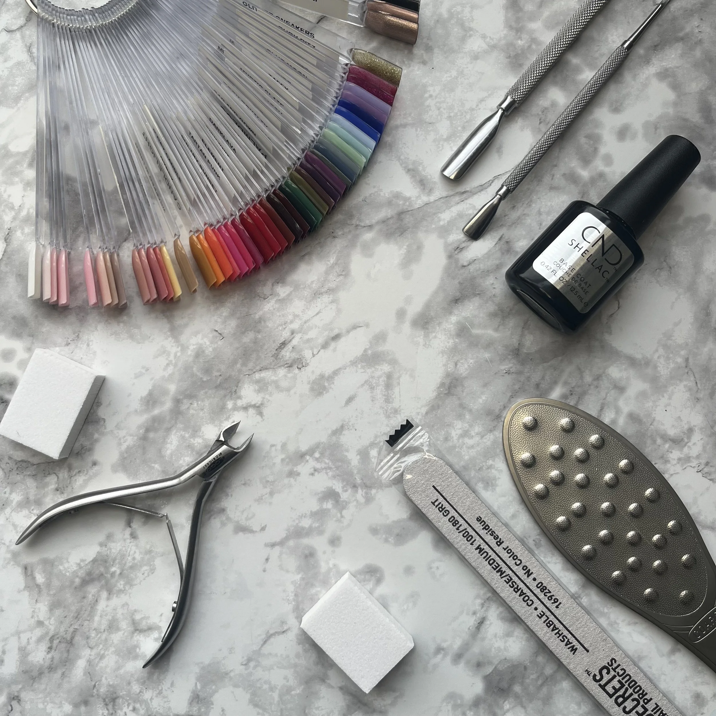 Nail polish color swatches, nail art tools, nail gel polish, nail cuticle nippers, nail buffer, manicure brush, and a foot file on a marble surface.