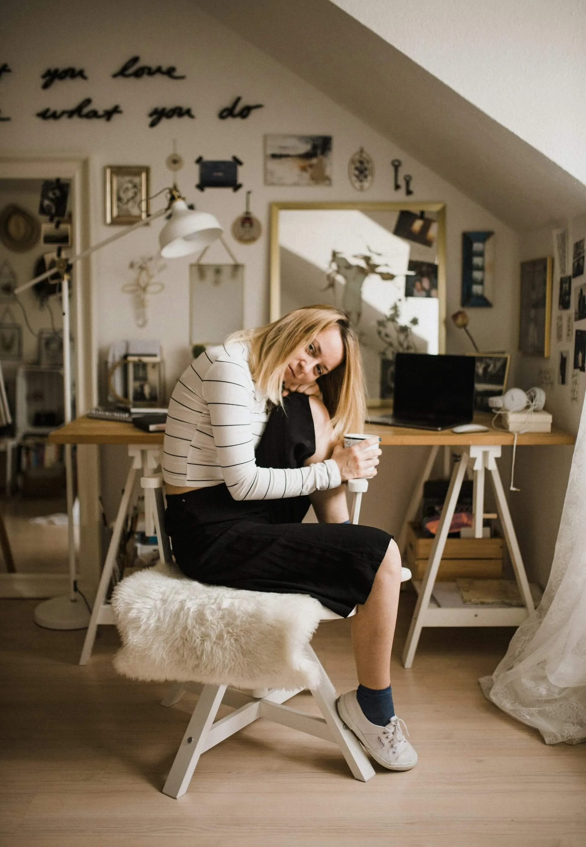A woman with blonde hair sitting on a white fur-covered stool in a cozy, decorated room, holding a mug, wearing a striped long-sleeve shirt and black skirt.