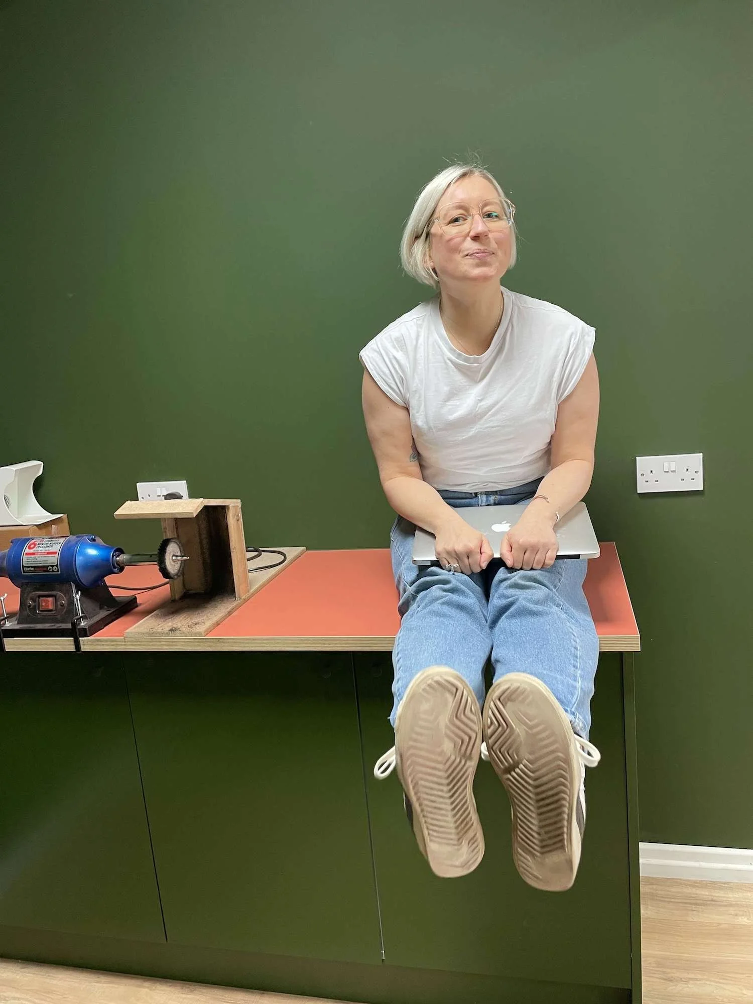 A woman with blonde hair and glasses sitting on a desk with her legs extended forward, holding a laptop, in front of a green wall.