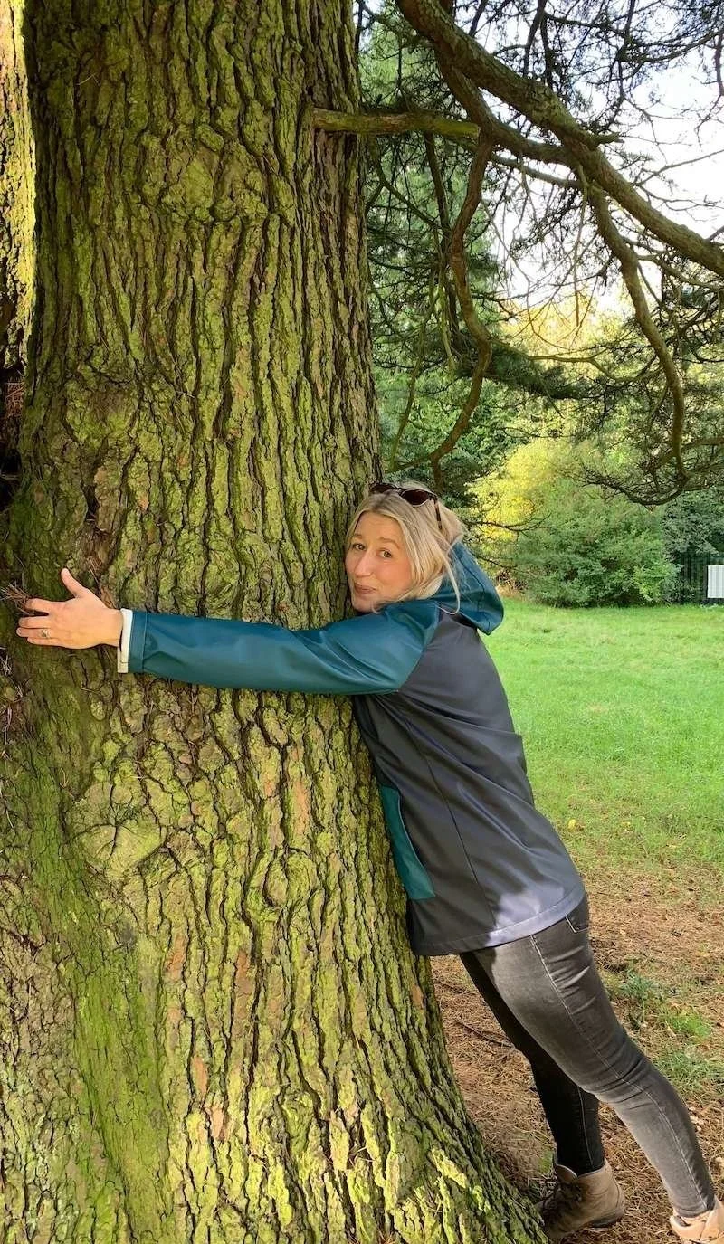 A woman with blonde hair hugging a large tree in a park, wearing a blue and black jacket, gray pants, and tan boots, with trees and grass in the background.