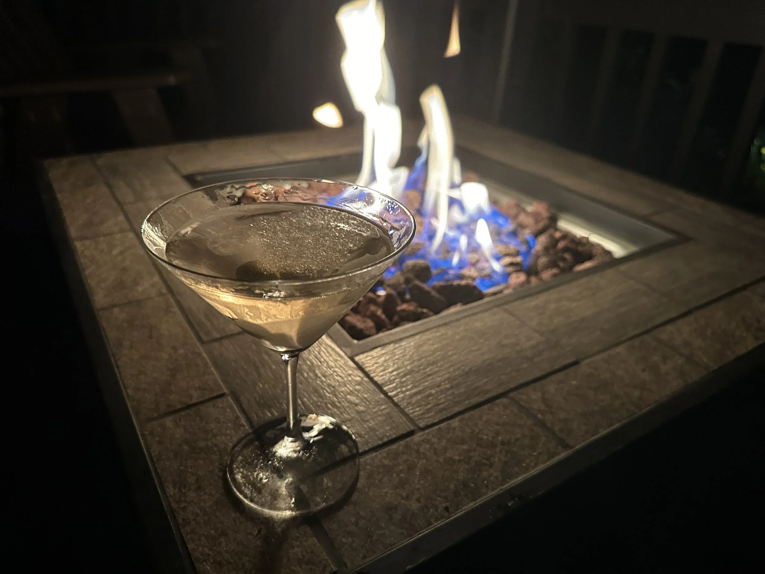 A perfectly crafted martini resting on the edge of the gas firepit, with warm flames flickering against the peaceful backdrop of the covered front deck.