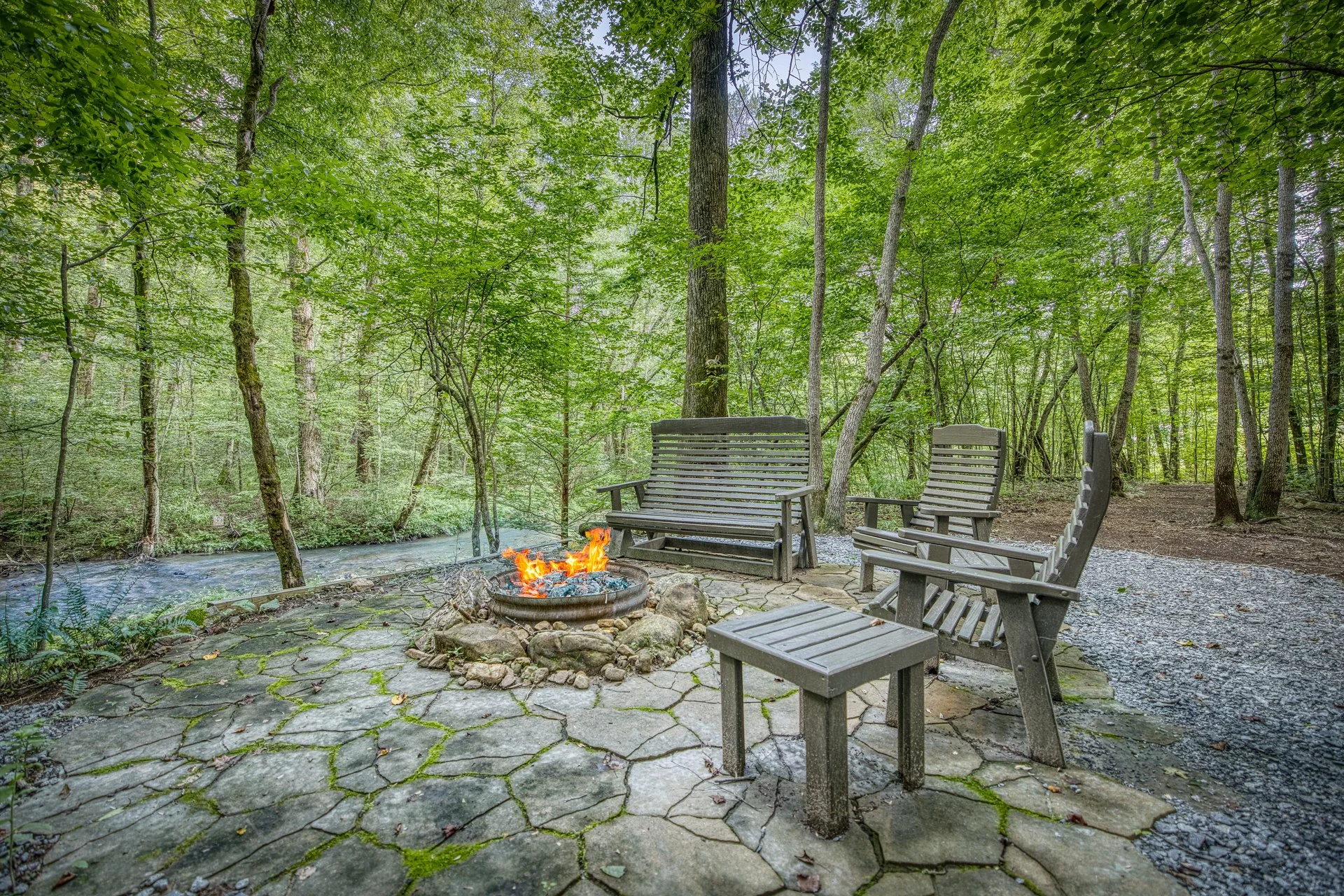 A cozy outdoor sitting area with four wooden chairs and a bench around a small fire pit, overlooking a river in a green forest in the North Georgia Mountains.