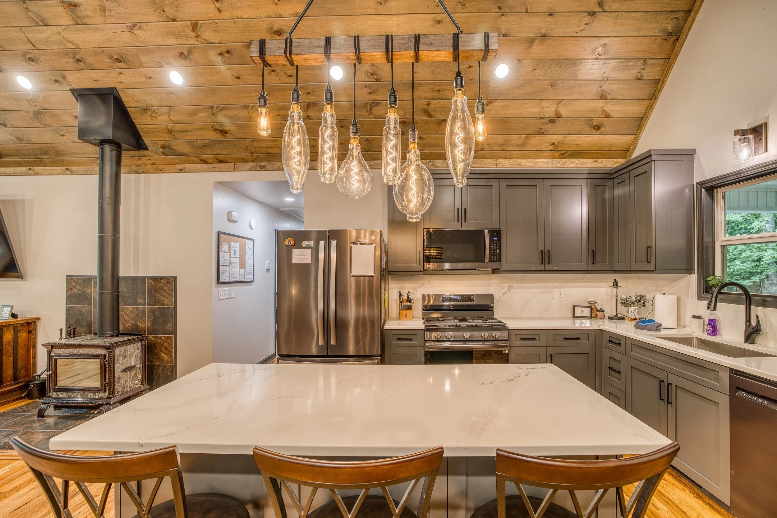 Modern kitchen with large quartz island, pendant lights, gas range, stainless appliances, and wood ceiling.