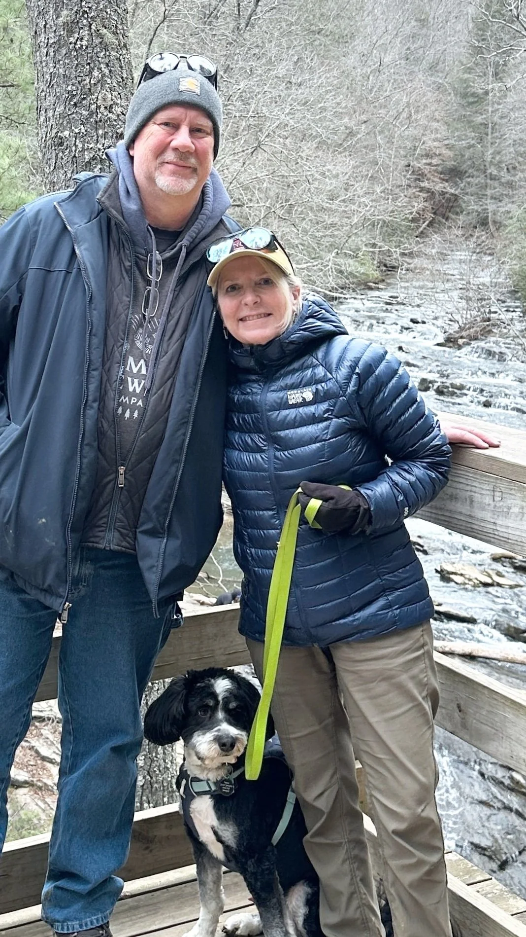 Hosts bundled up in early spring attire posing on a deck overlooking a cascading creek by Carters Lake, included in the picture is their mini bernedoodle.