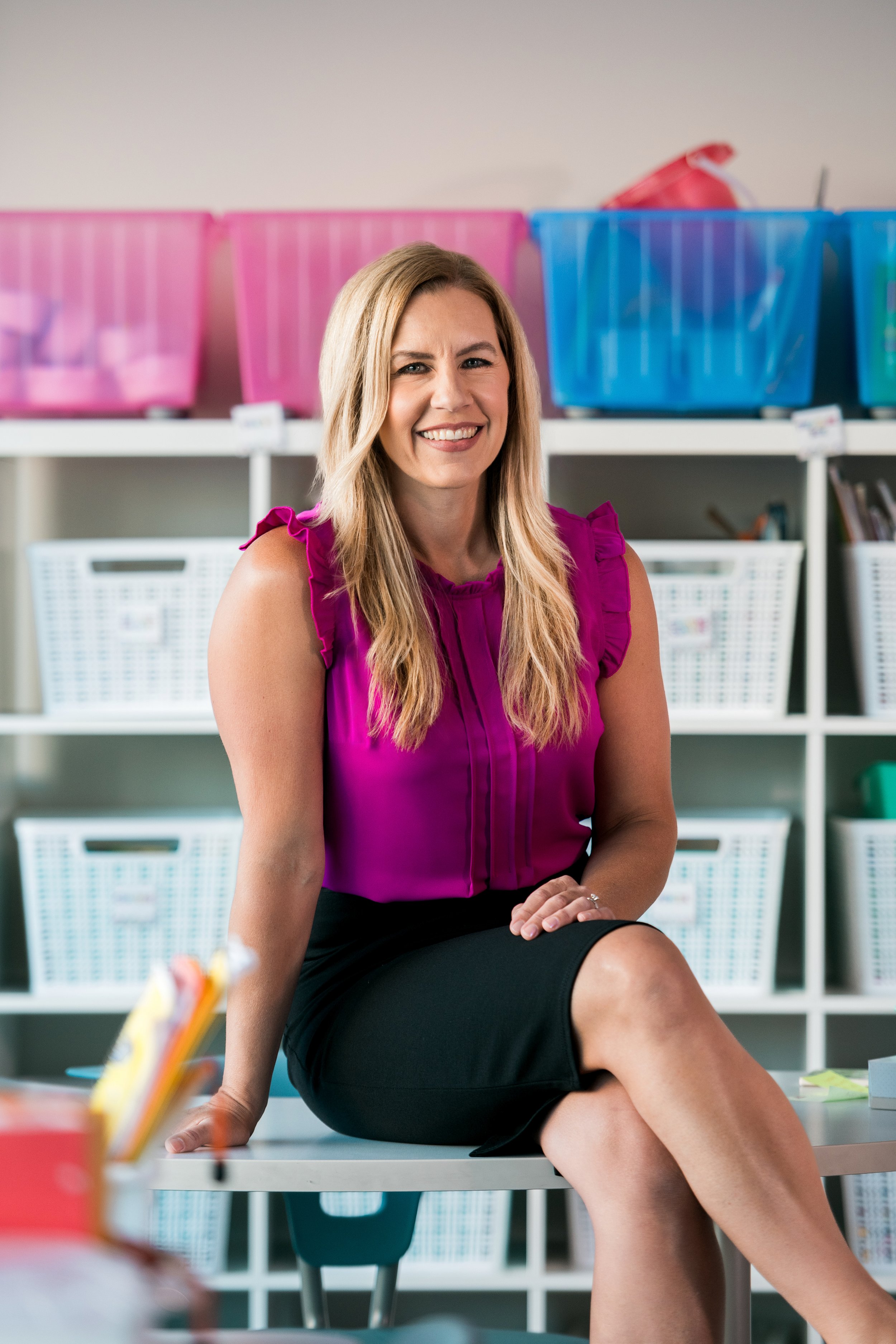 Teacher sitting on a table in her classroom facing the camera with classroom materials in the background.