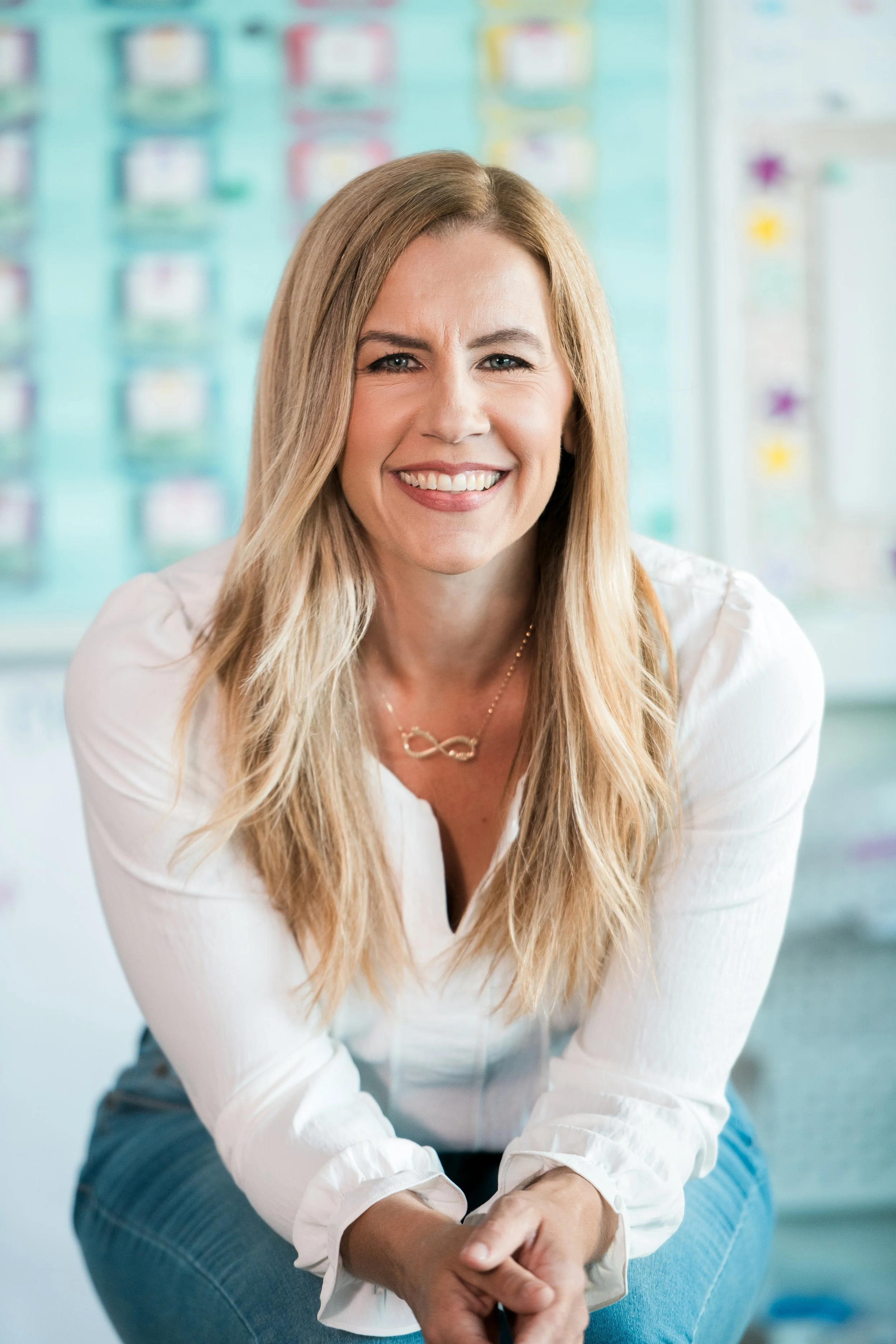 Teacher sitting in her classroom leaned over and smiling at the camera.