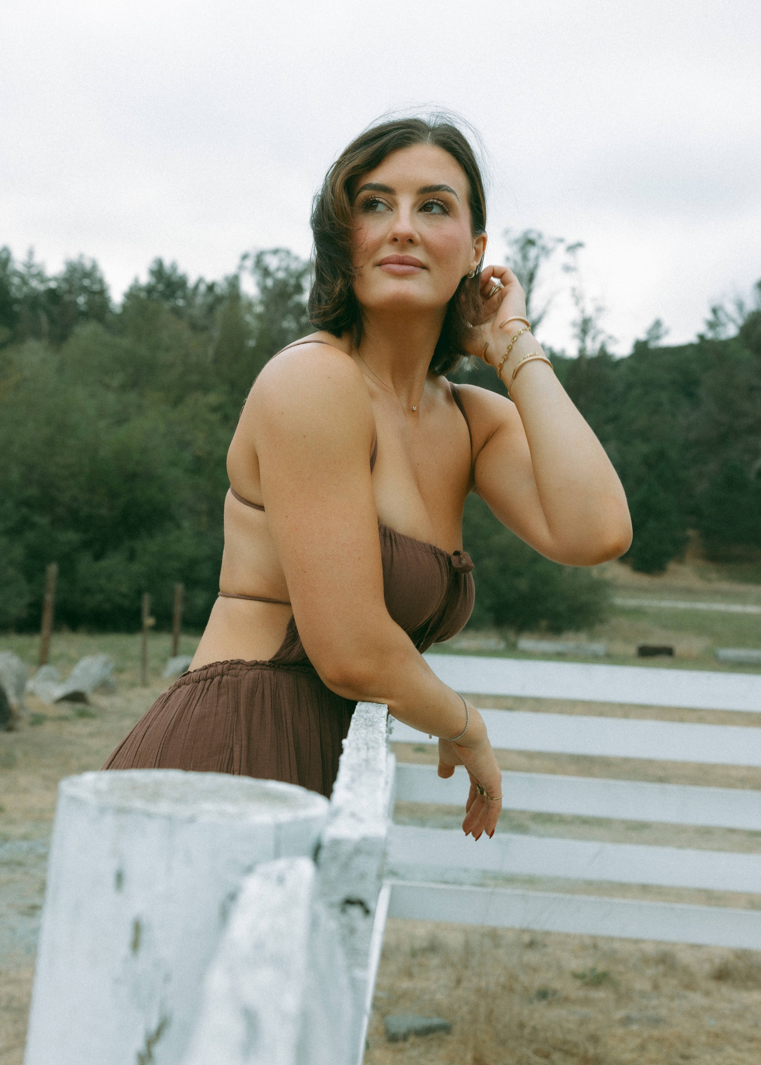 A woman with shoulder-length dark hair wearing a brown sleeveless dress posed outdoors leaning on a white fence, with a backdrop of trees and a cloudy sky.