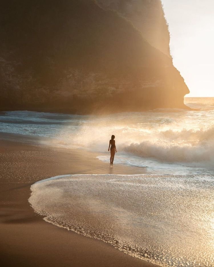 A person standing at the shoreline on a beach, with cliffs and the setting sun in the background.