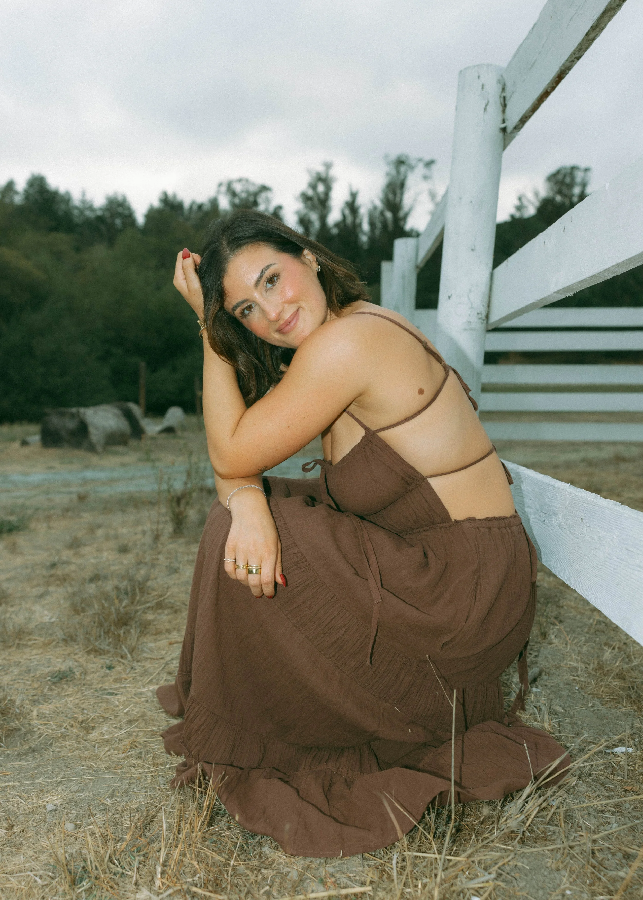 A woman in a brown dress sits outdoors next to a white wooden fence, with her head resting on her arm and smiling at the camera, in a natural setting with trees in the background.