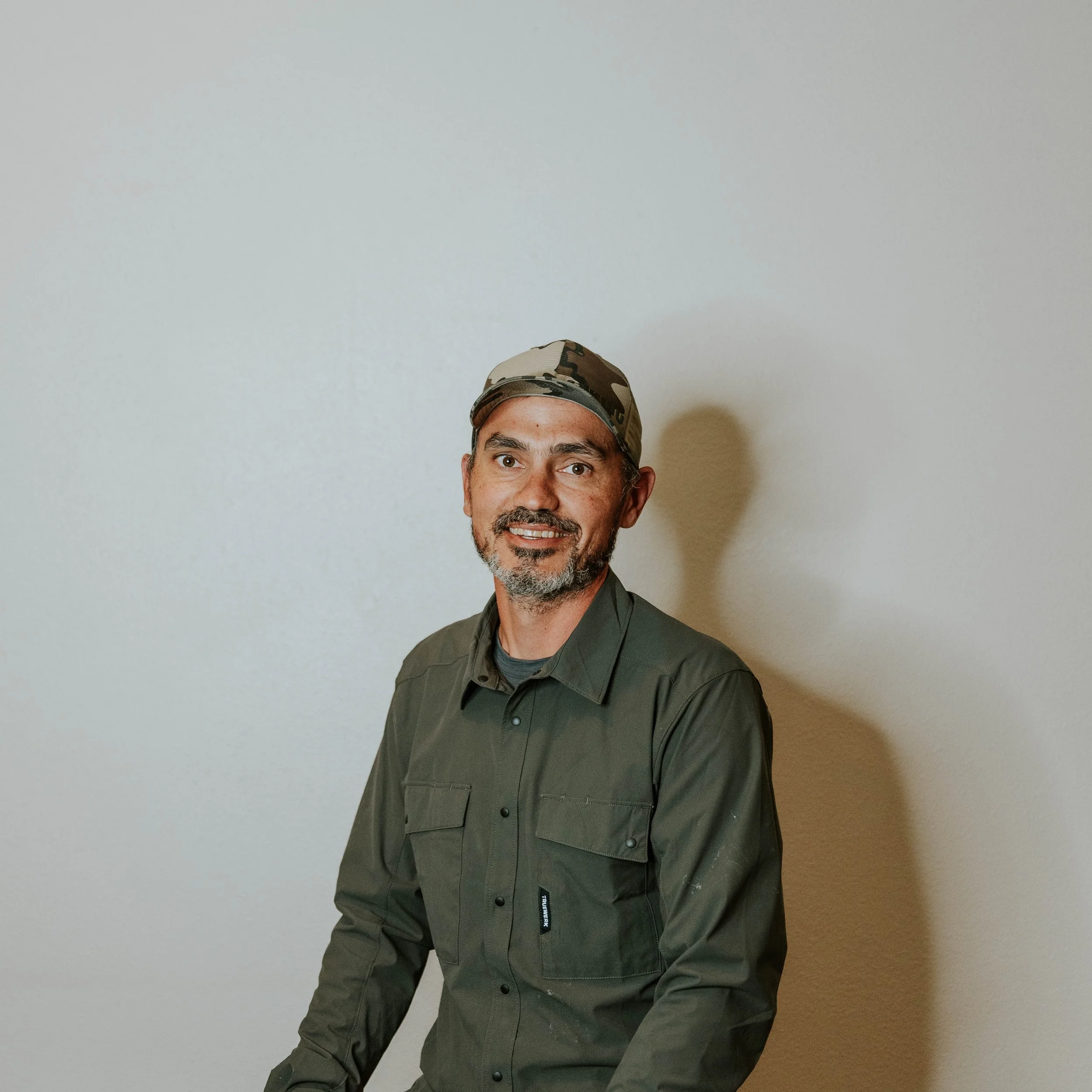 Person wearing a brown cowboy hat and a hoodie, smiling under a blue sky with clouds and trees in the background.