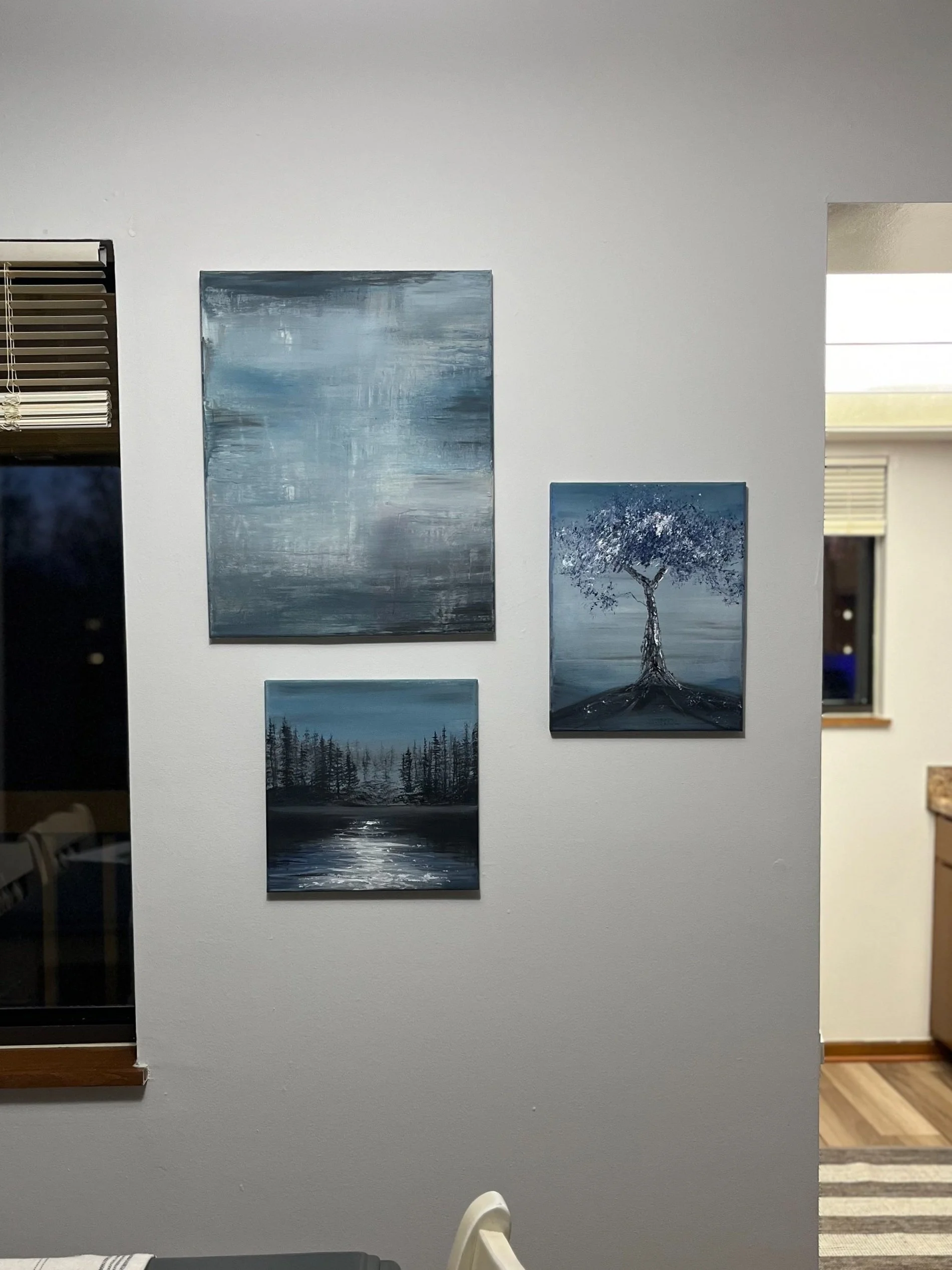 Three paintings with blue and gray tones on a white wall in a home interior.