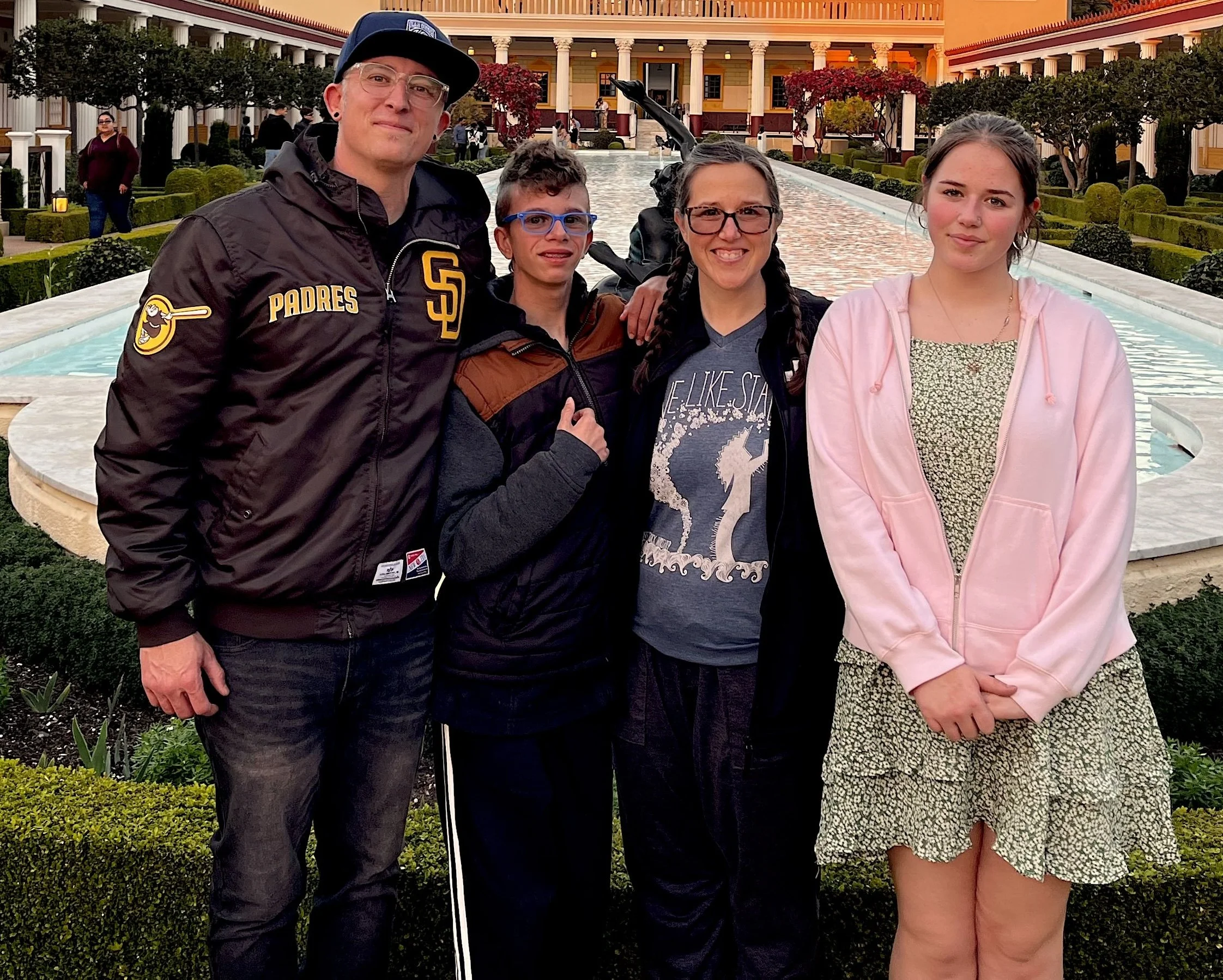 A group of five people standing together outside in front of a decorative water fountain and a building with columns, with some trees and shrubs around them. They are smiling and posing for the photo.