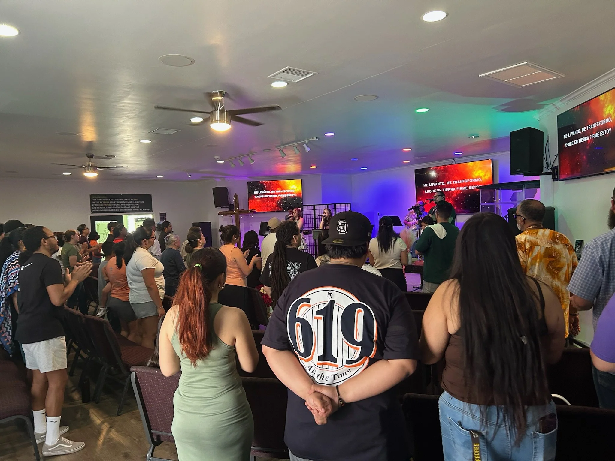 People gathered in a church or worship space, some standing and some clapping, while a band performs on stage with screens displaying lyrics.