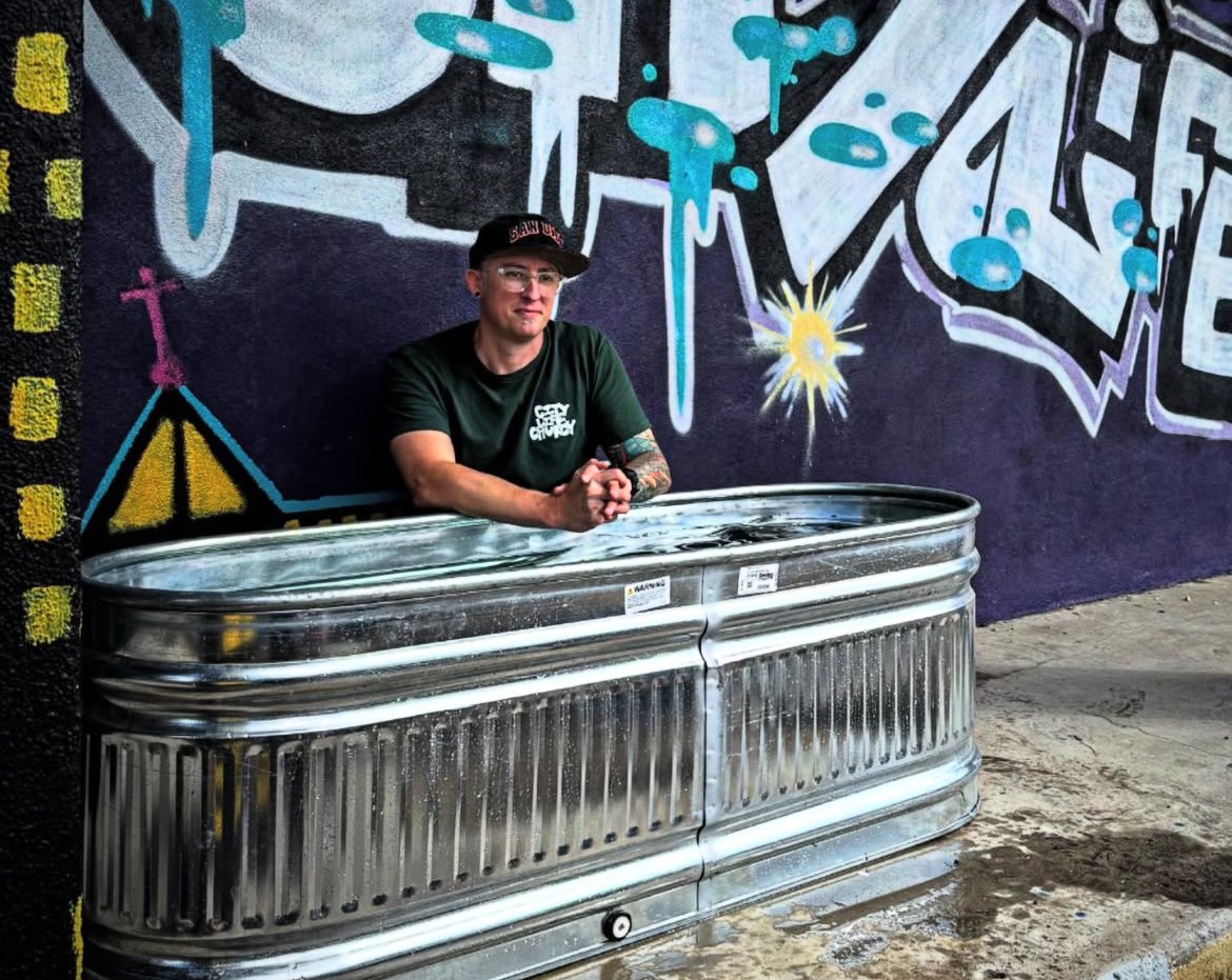 A person standing in a metal tub filled with water in front of a colorful graffiti wall.