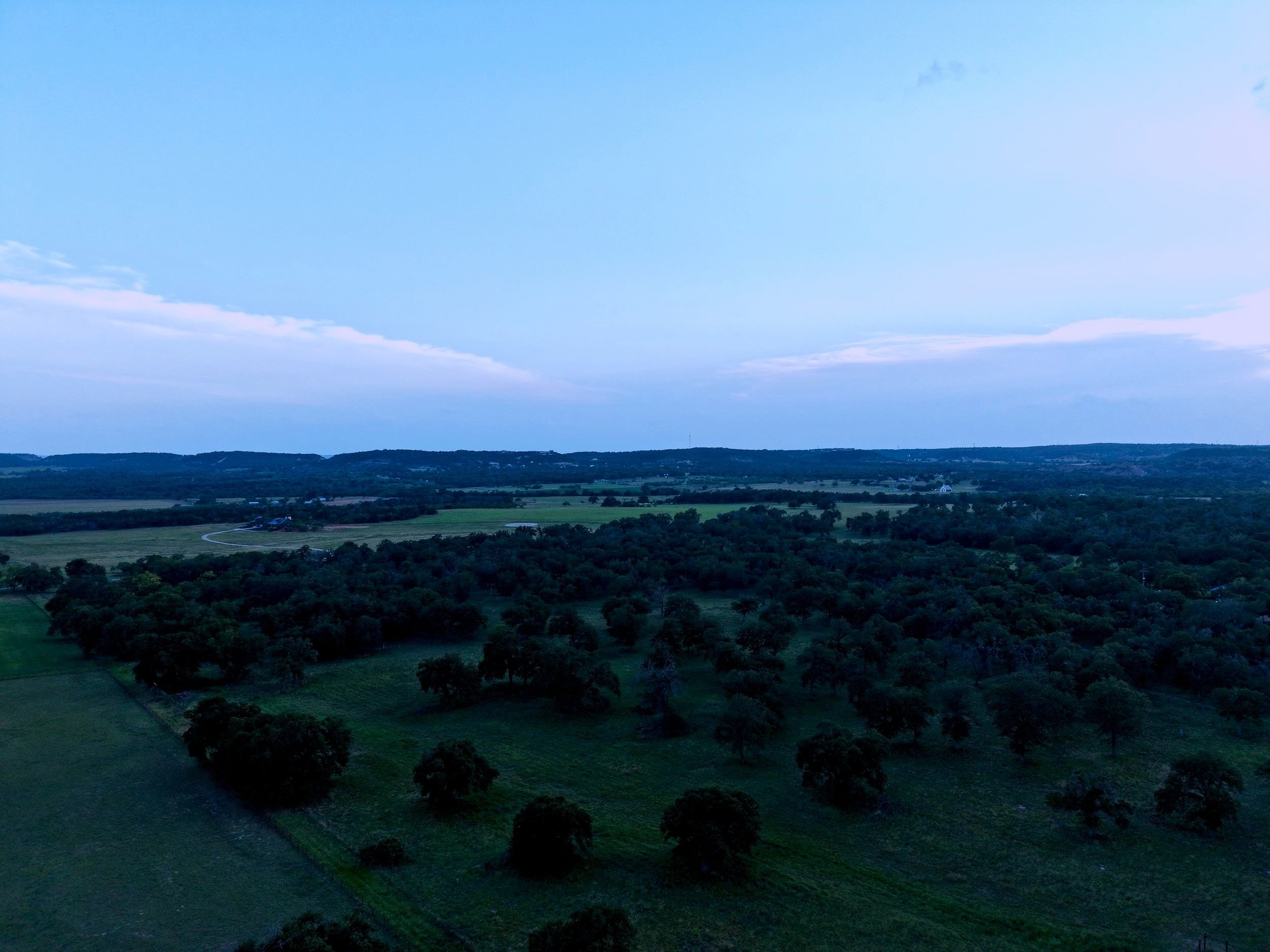 Aerial view of a green landscape with fields, trees, and distant hills under a partly cloudy sky.