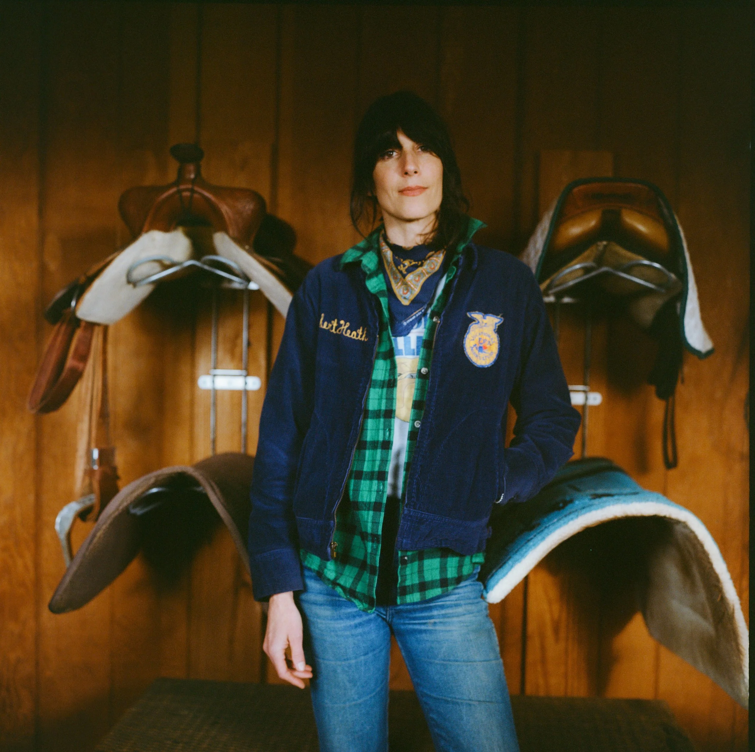 A woman with dark hair and a serious expression stands in front of a wooden wall with two equestrian saddles hanging behind her. She wears a navy blue jacket with embroidered logo and the words 'Lettheath', a green plaid shirt, and jeans.