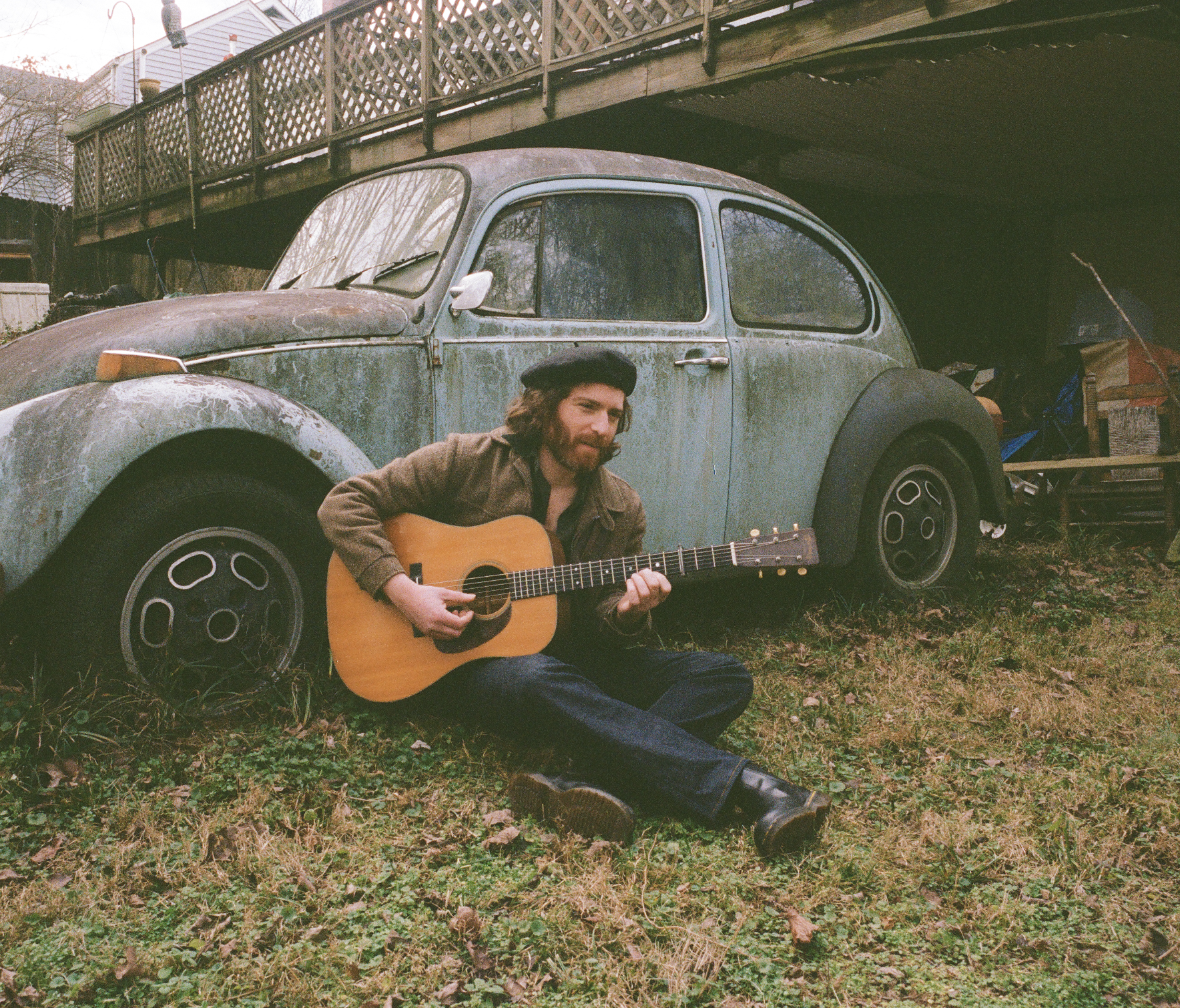 A man with long hair and beard, wearing a black beret and a brown jacket, sitting on grass and playing an acoustic guitar beside an old, weathered Volkswagen Beetle in a backyard with a wooden fence and shed.
