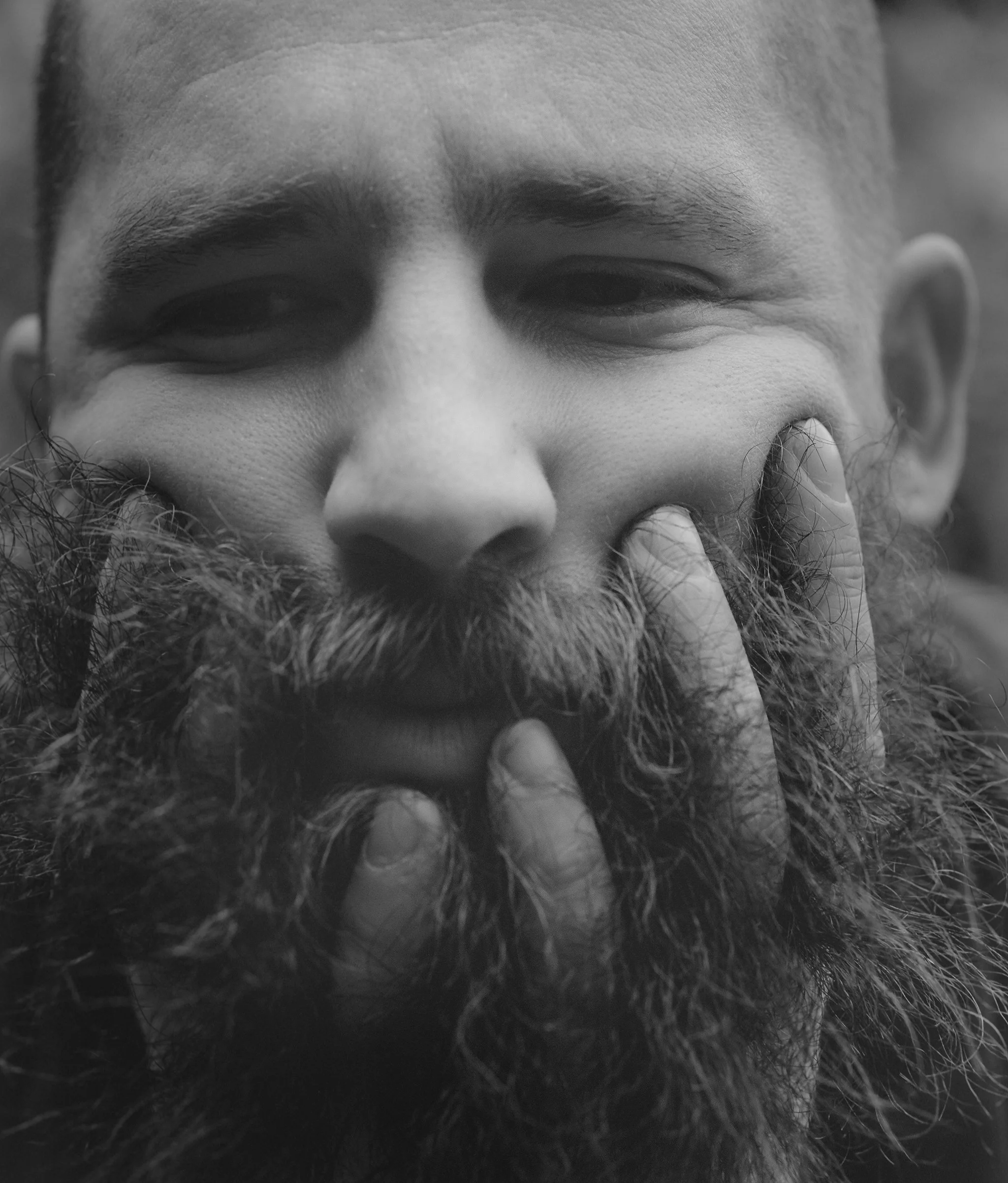 Close-up of a man with a thick beard and mustache, holding his hand up to his face and making a peace sign over his lips, in black and white.