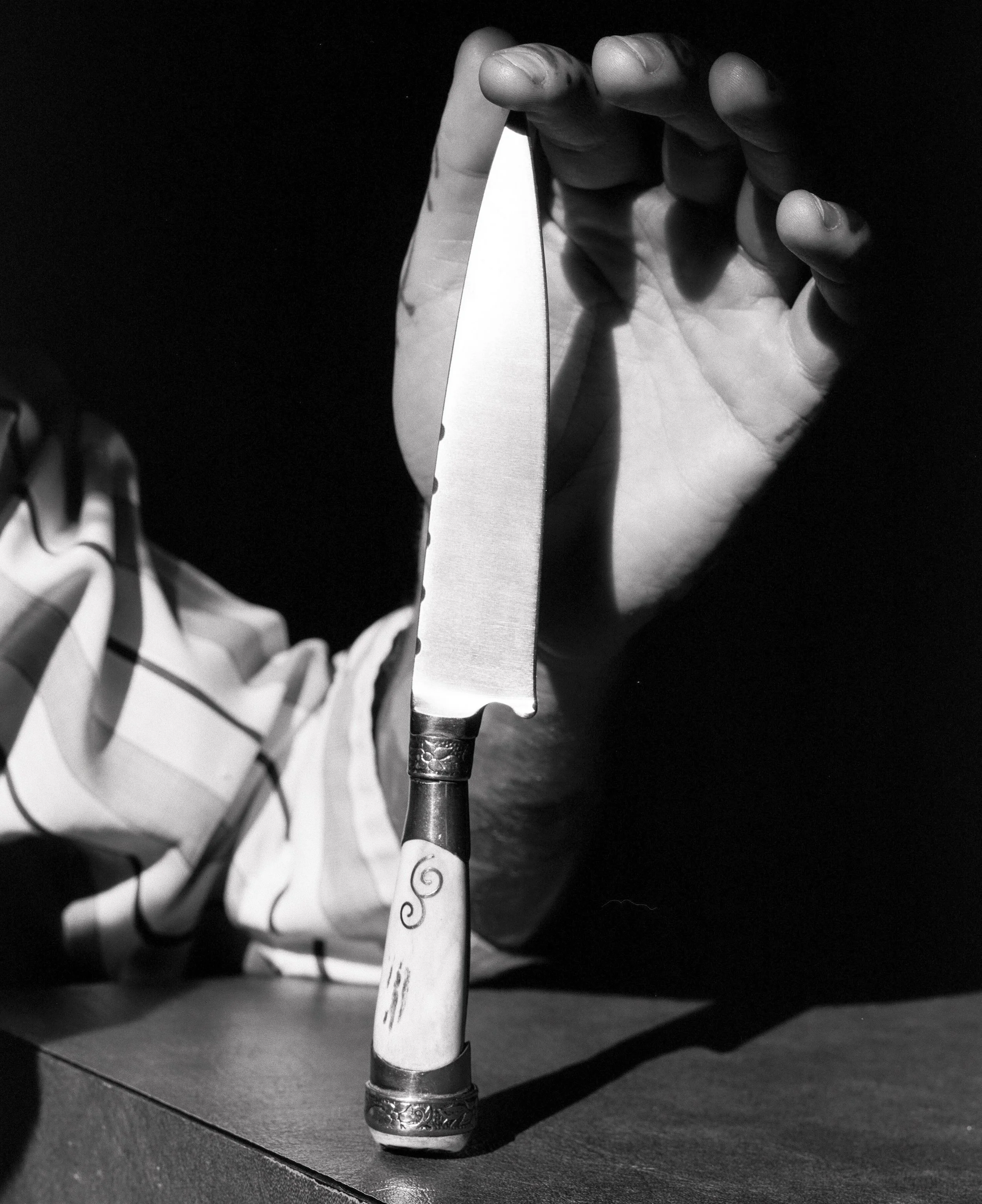 A black and white photo of a hand holding a large knife with a decorated handle, above a wooden surface, with a dark background.