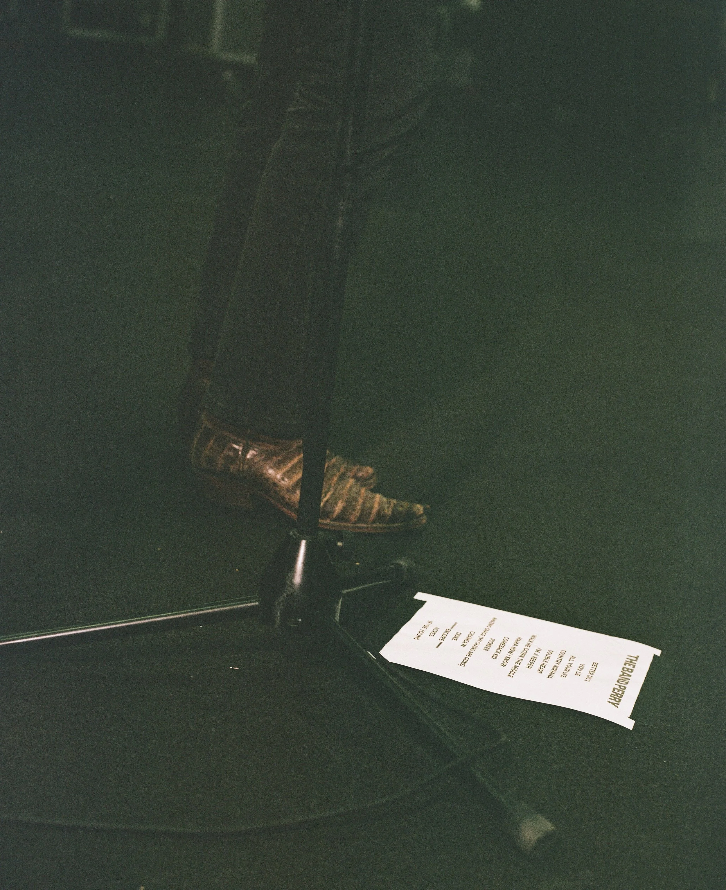 Close-up of a person's legs and feet wearing leather shoes and dark pants, standing on a black stage with a microphone stand and music playlist on the floor.