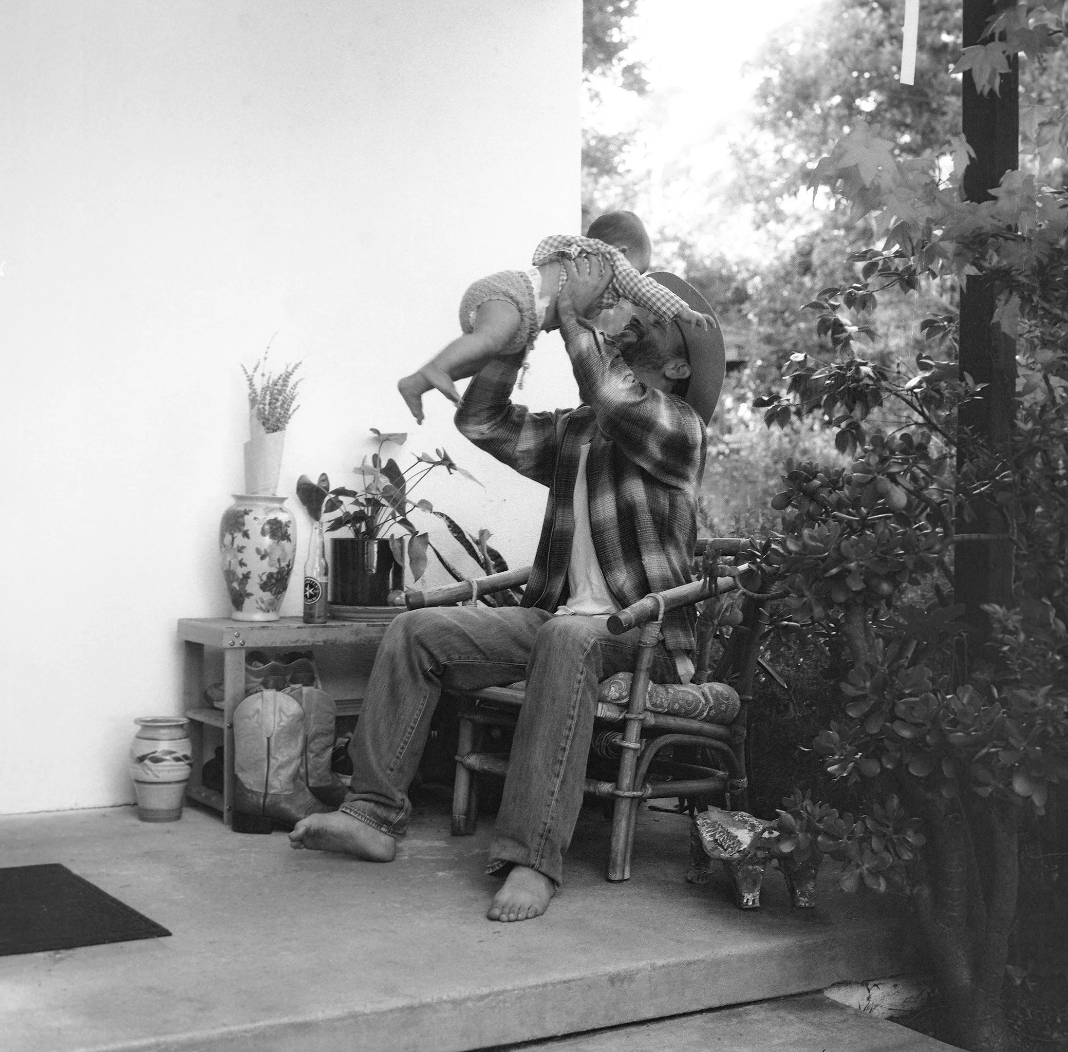 A man sitting in a wooden chair, lifting a young child towards him outdoors, with plants and trees in the background, in a black and white photo.