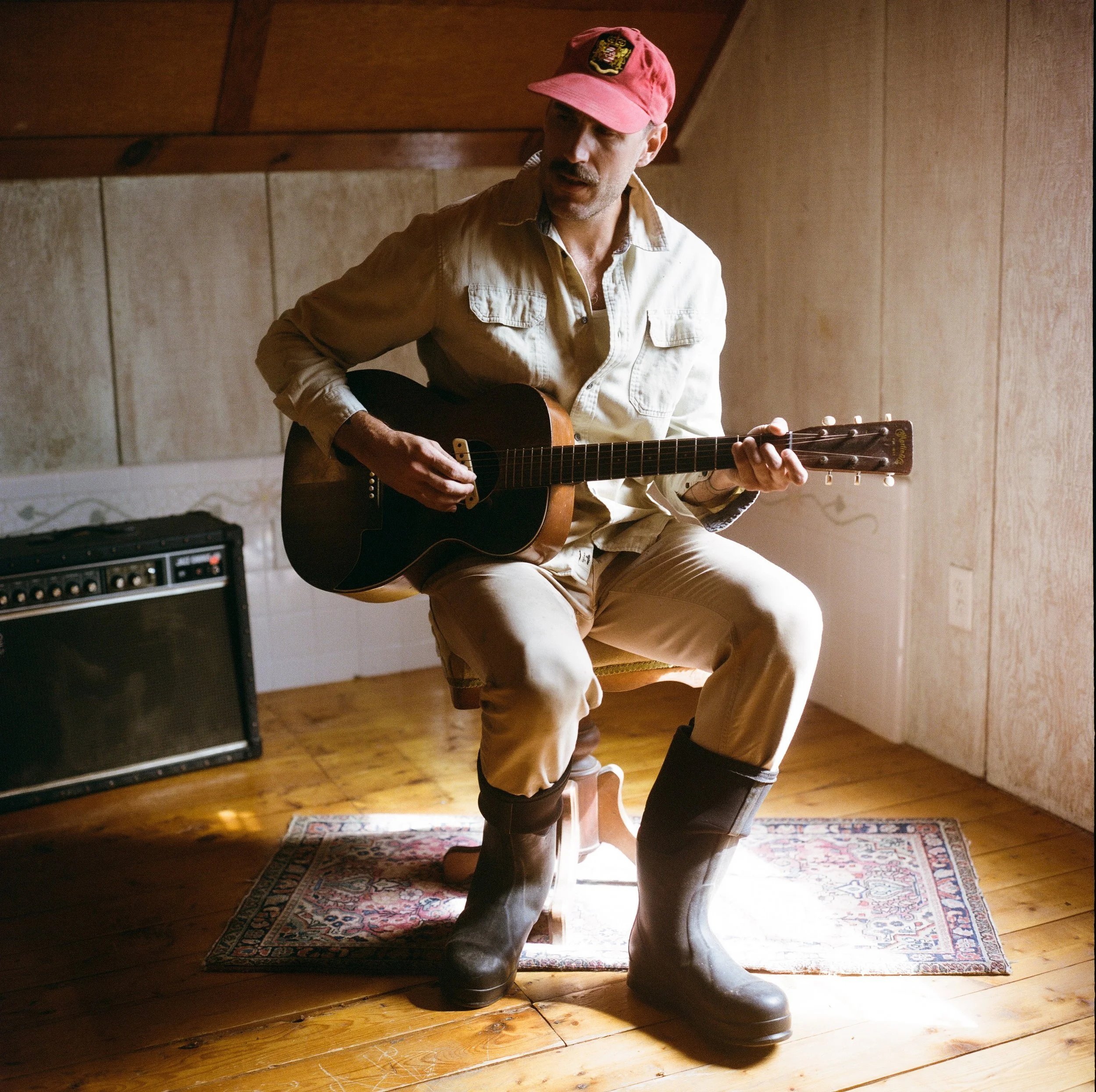 A man wearing a red baseball cap, beige shirt, and beige pants, sitting on a wooden chair, playing an acoustic guitar. An amplifier is on the floor beside him, and a small rug is under his feet. The setting appears to be a cozy, wood-paneled room.