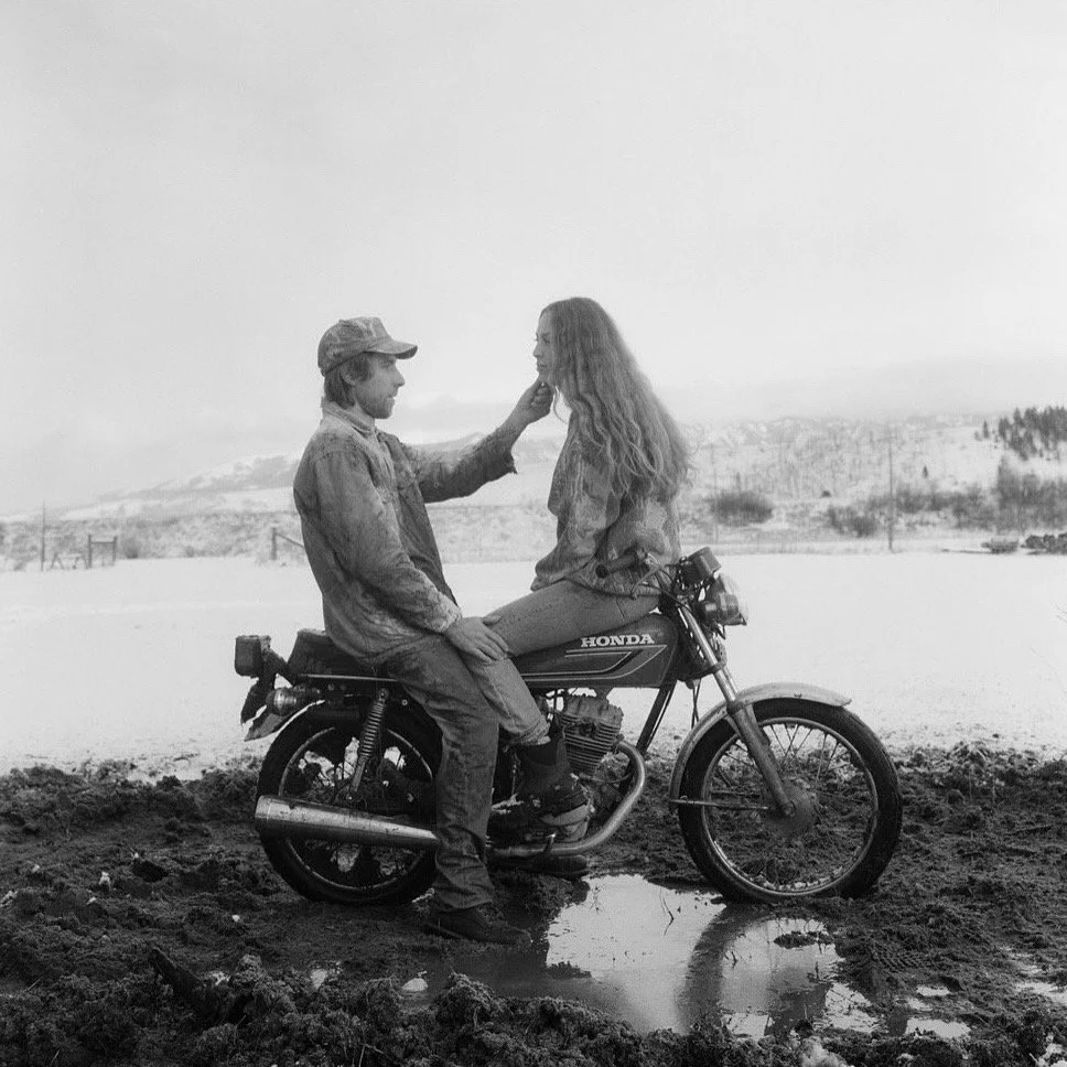 A man and woman sitting on a motorcycle outdoors in a muddy area, with snow-covered hills in the background. The man is touching the woman's face as they look at each other.