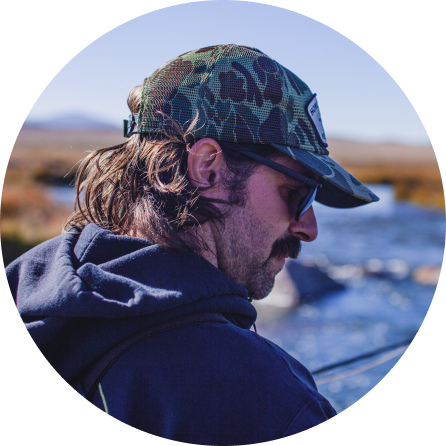 A man with long hair, sunglasses, and a camouflage baseball cap, wearing a navy hoodie, standing outdoors near a river.