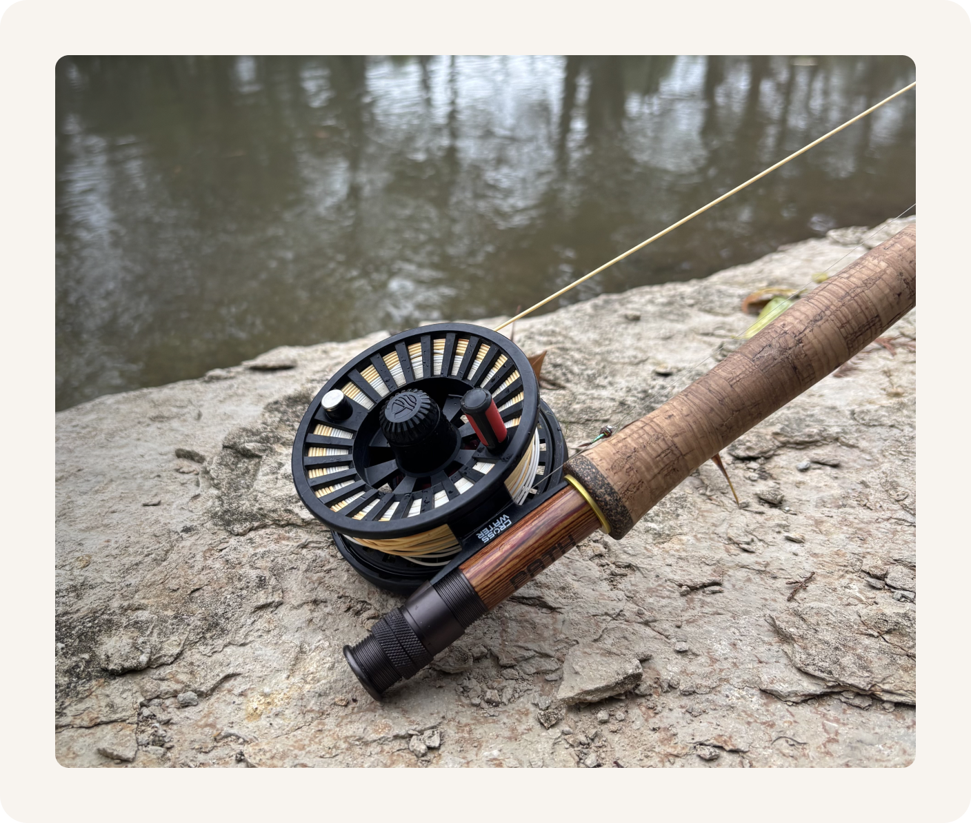 A fly fishing rod with a reel resting on a rocky shoreline next to a body of water.