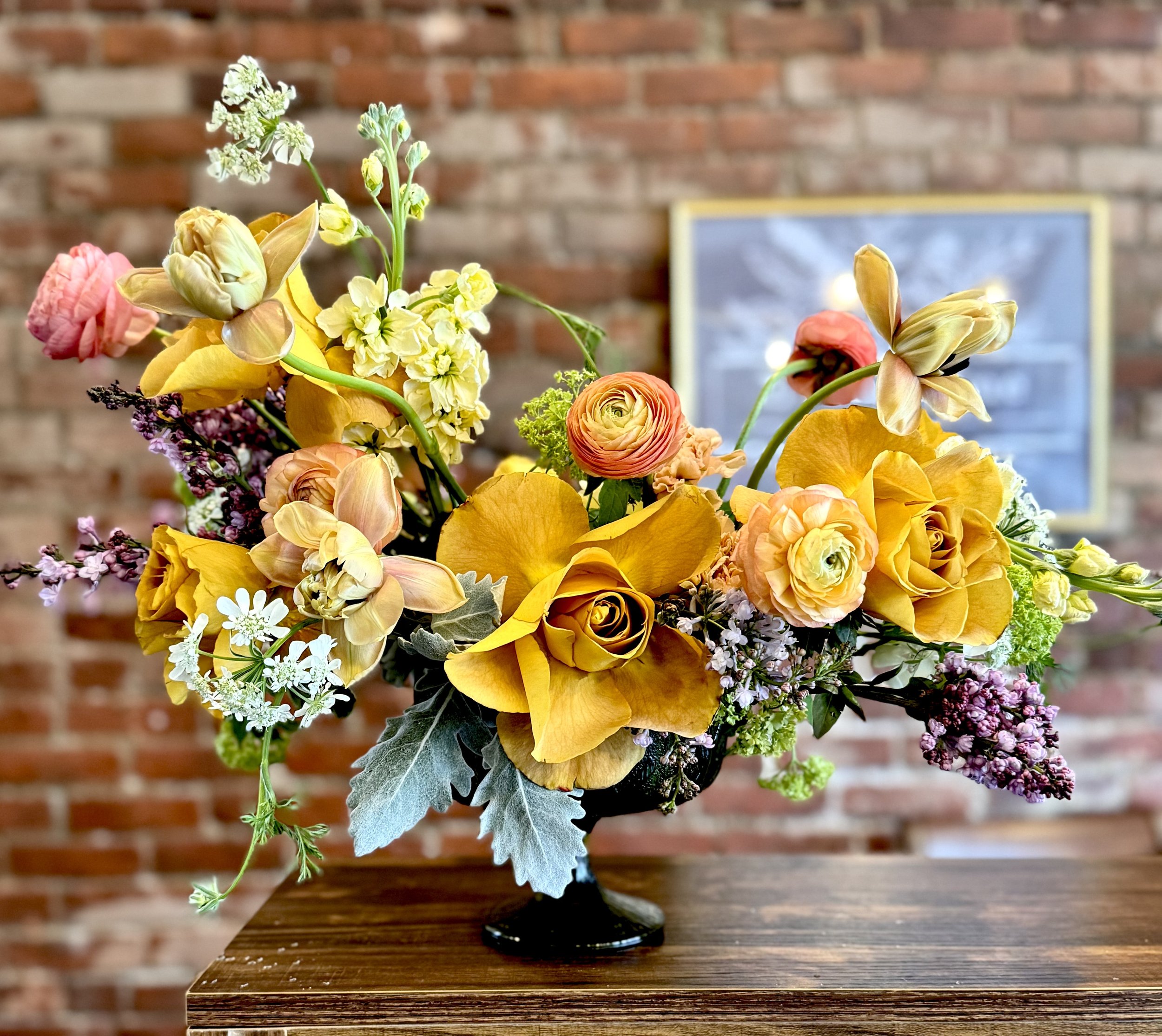 Floral arrangement with yellow, orange, and pink flowers in a vase on a wooden table, brick wall background.