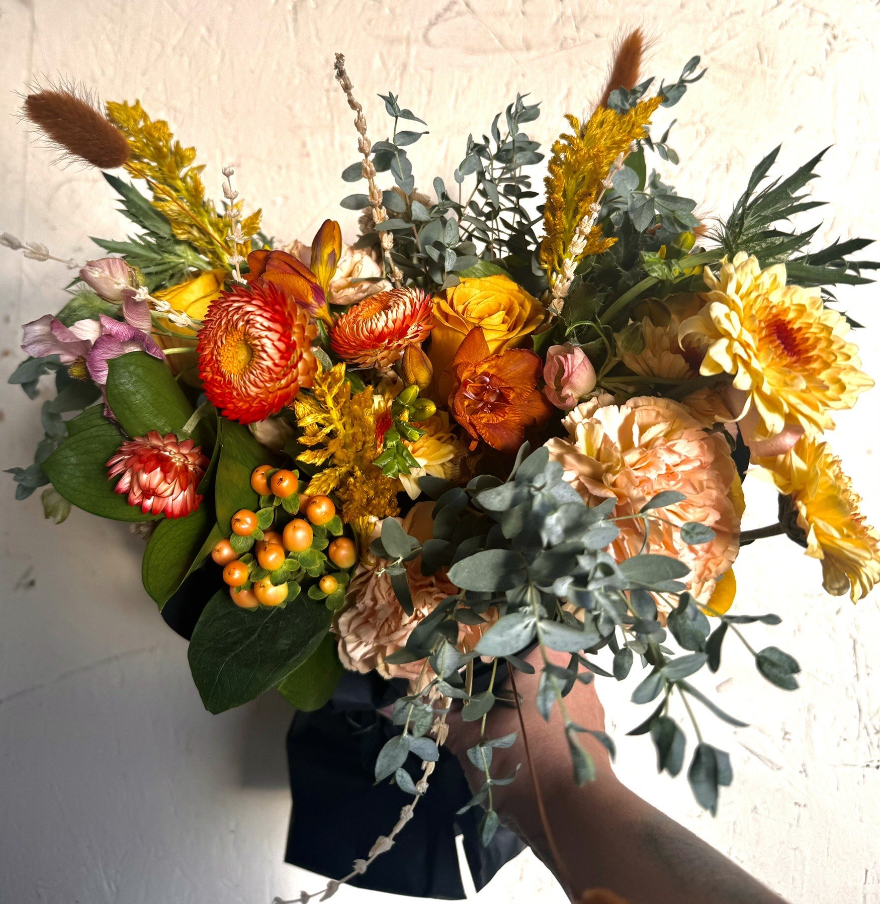 Hand holding a colorful bouquet with orange, yellow, red, and pink flowers, green foliage, and mixed textures.