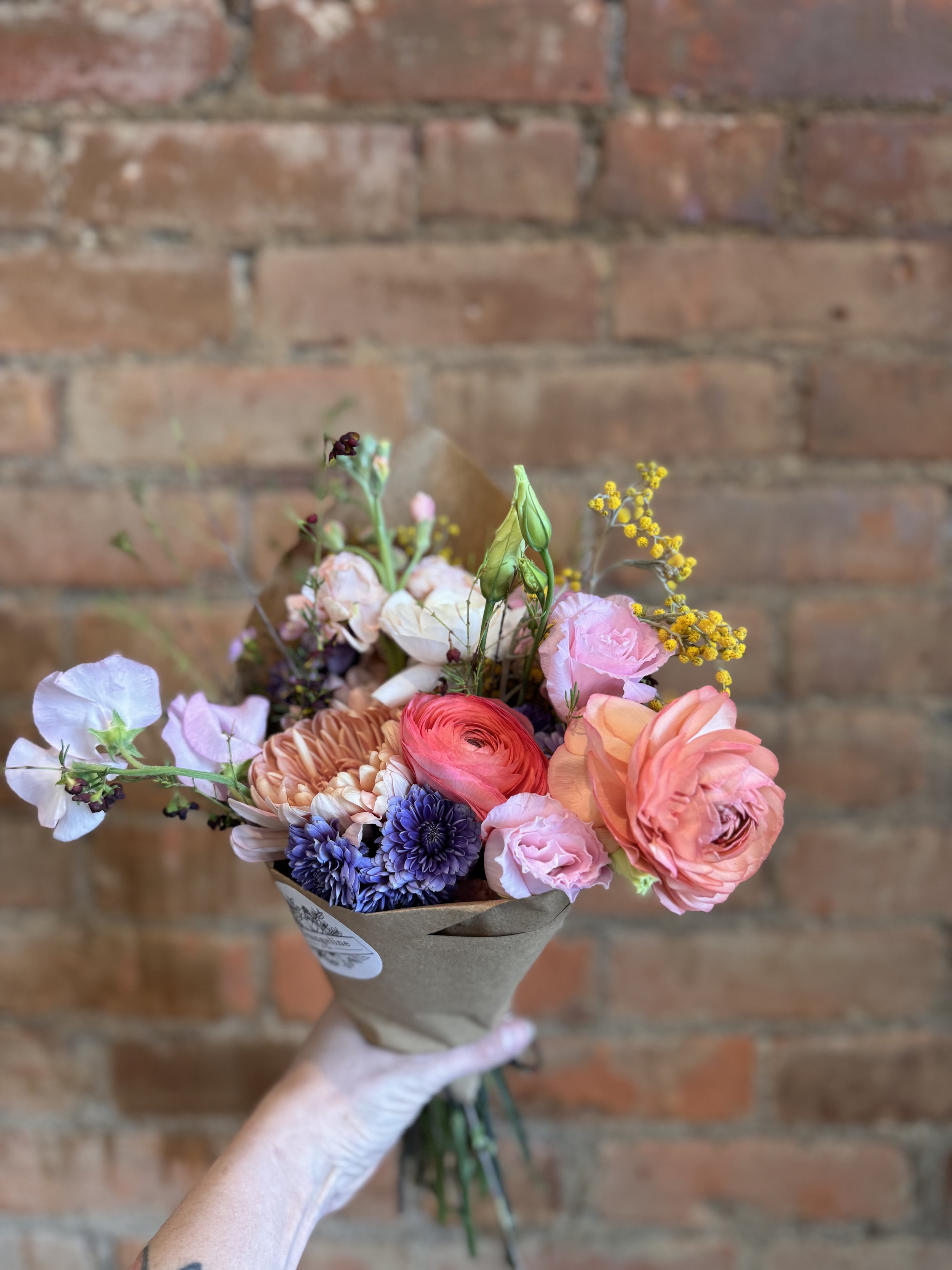 A hand holding a bouquet of colorful flowers, including pink roses, orange and coral ranunculus, purple and pink flowers, against a brick wall background.