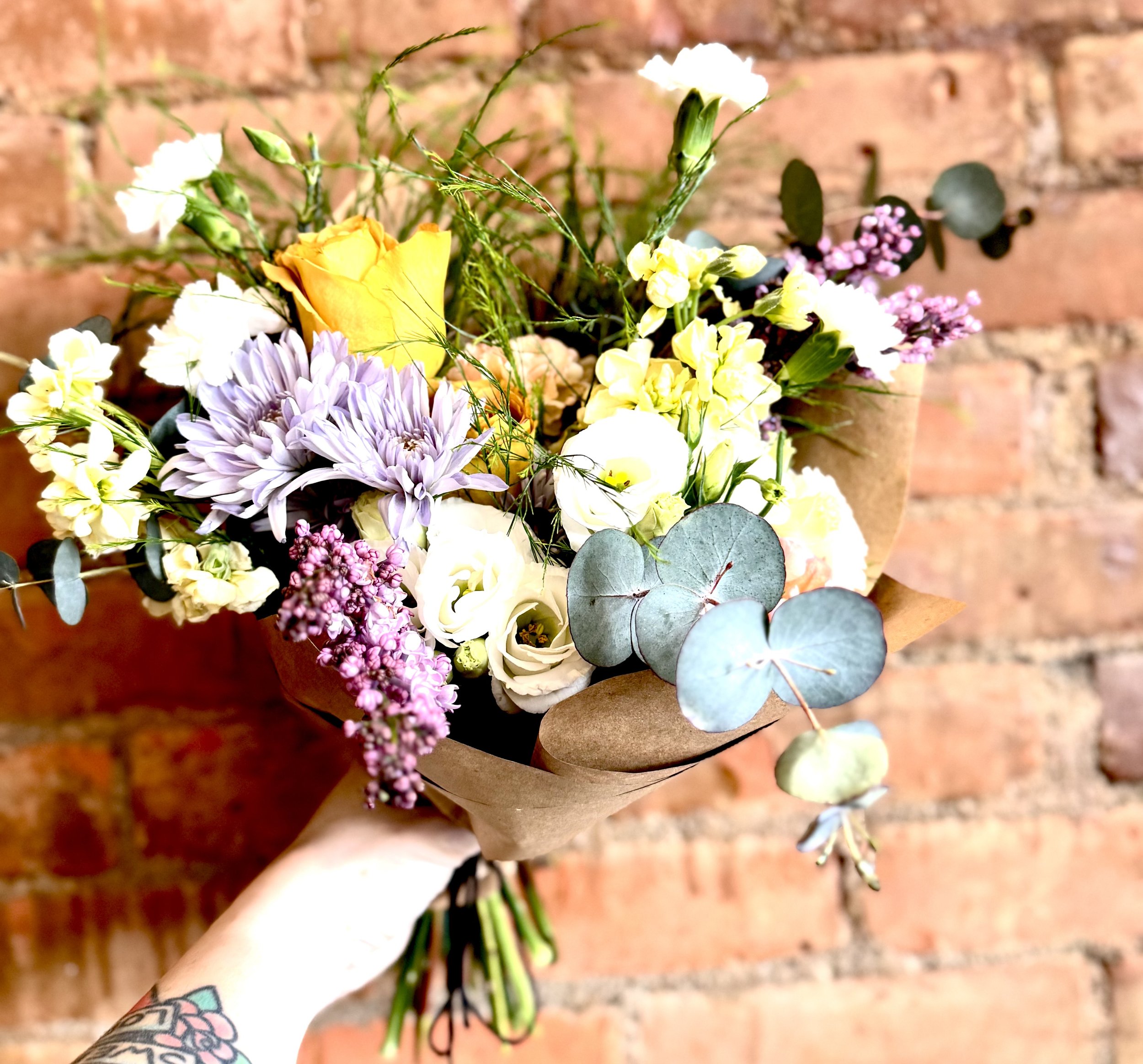 Colorful flower bouquet with roses, eucalyptus, and mixed blooms against brick wall.