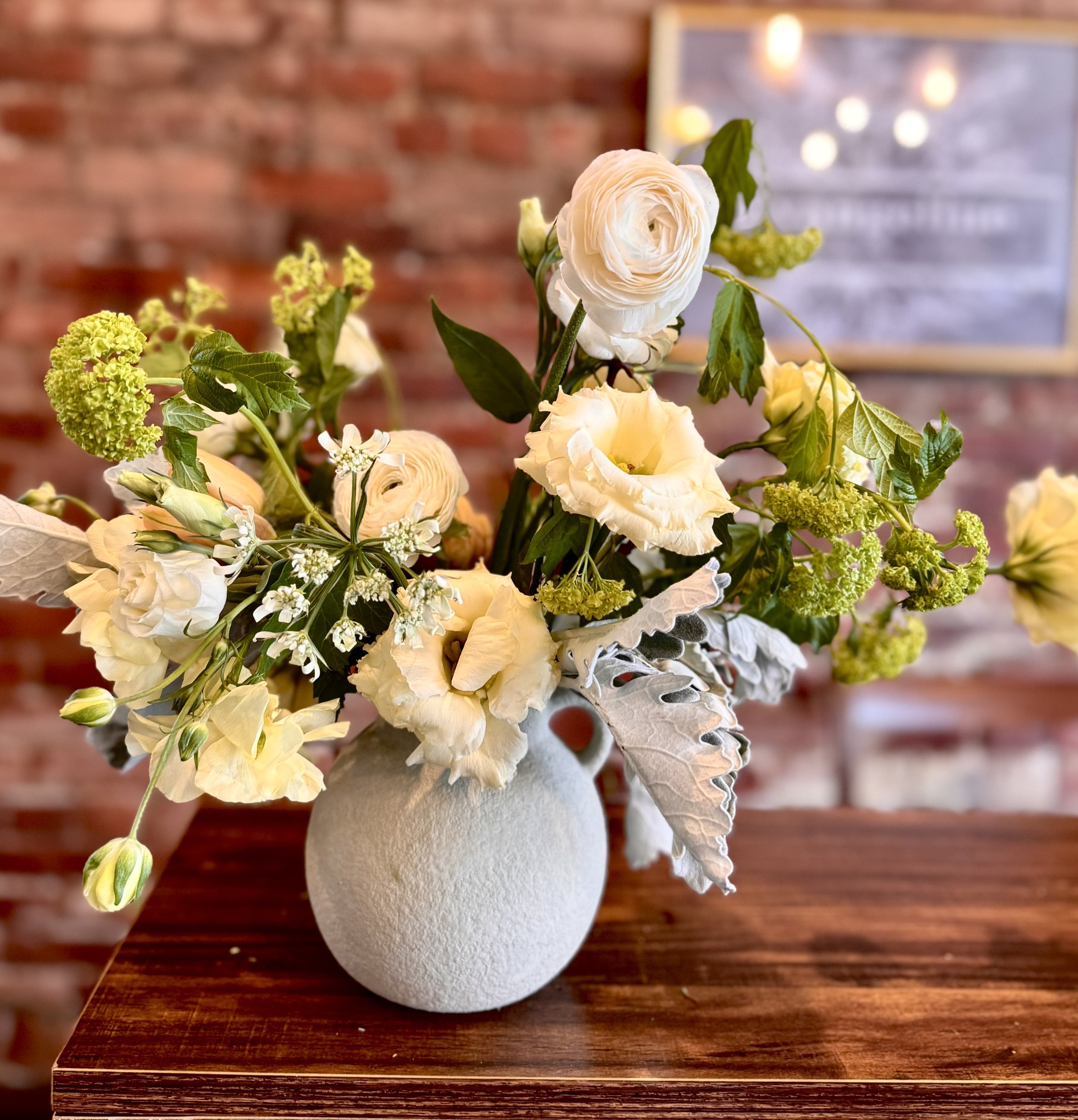 Bouquet of white and green flowers in a gray vase on a wooden table with a brick wall background.