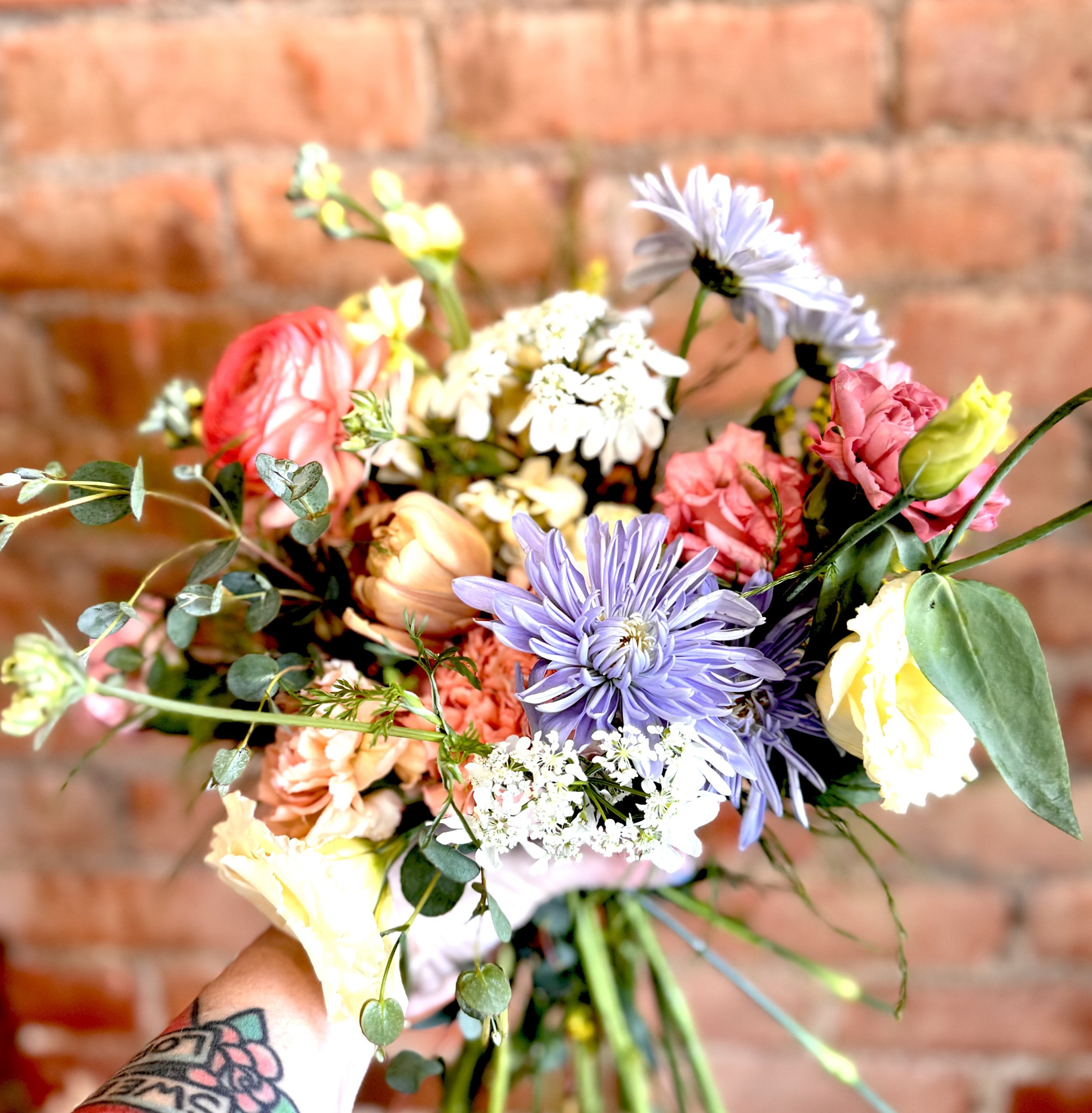 Bouquet of mixed flowers with purple, pink, and yellow blooms against a brick wall backdrop.