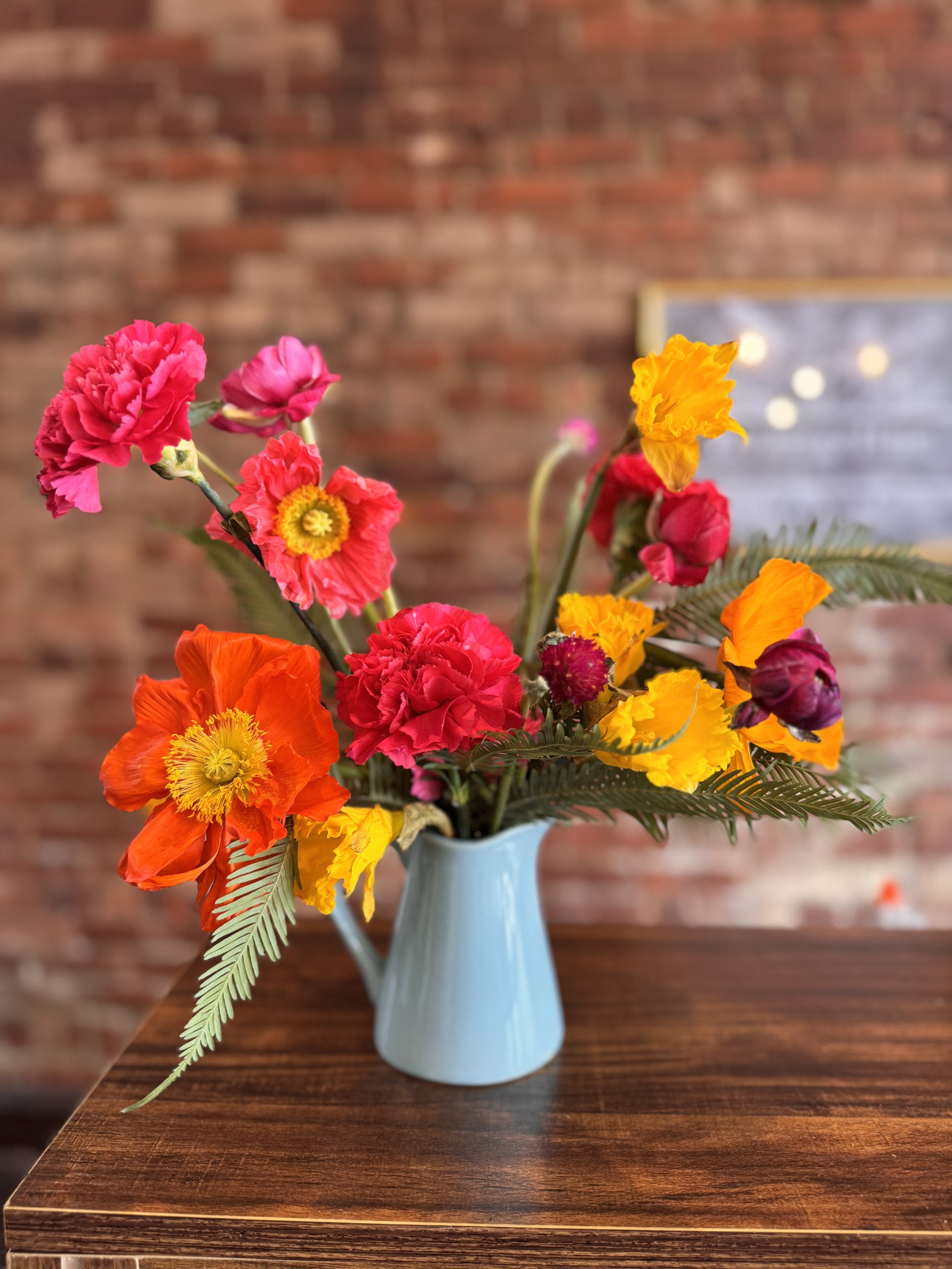 Colorful flower arrangement in a light blue pitcher on a wooden table, with a blurred brick wall background.
